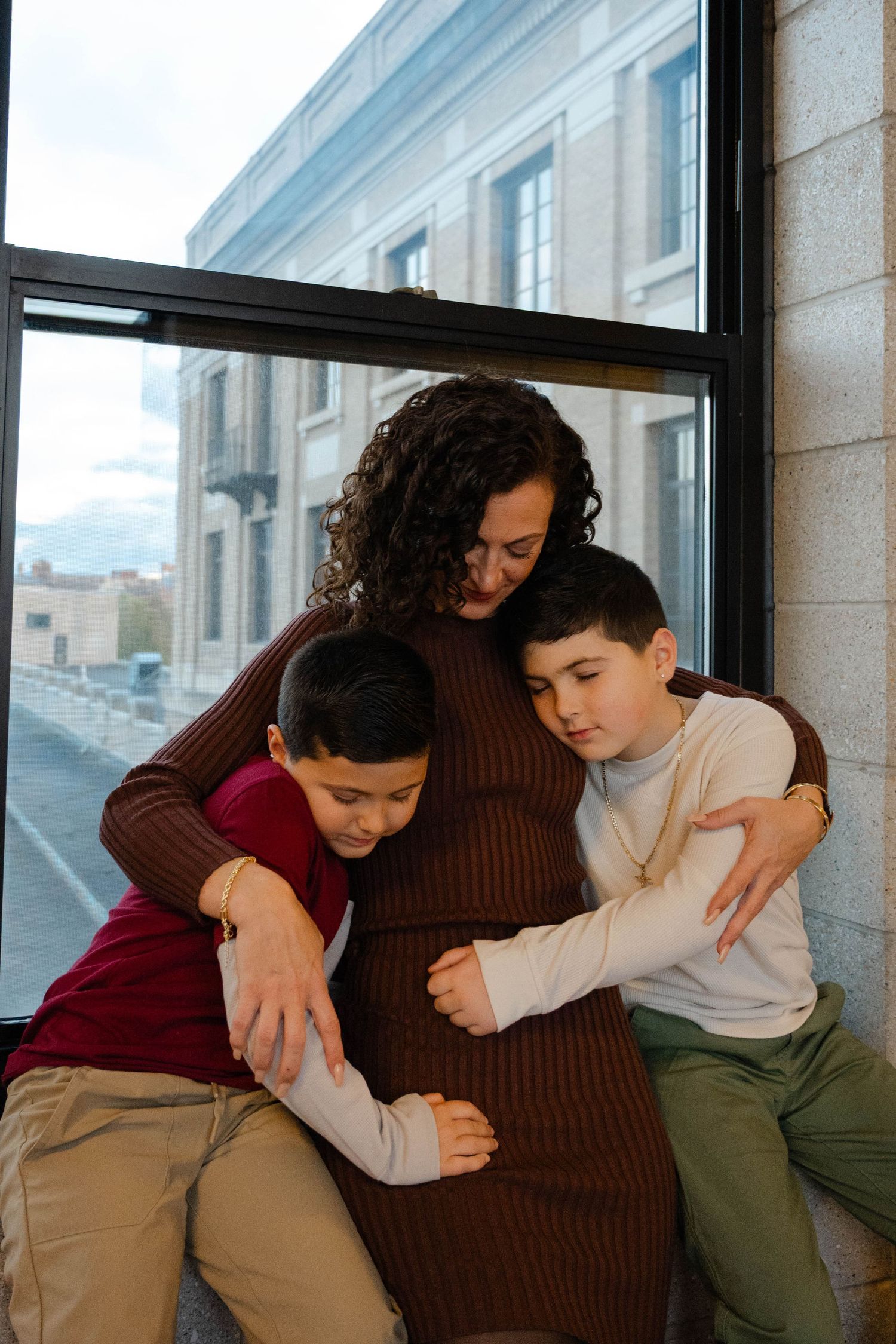 Two boys embrace their pregnant mother's belly while sitting by a window in a modern building.