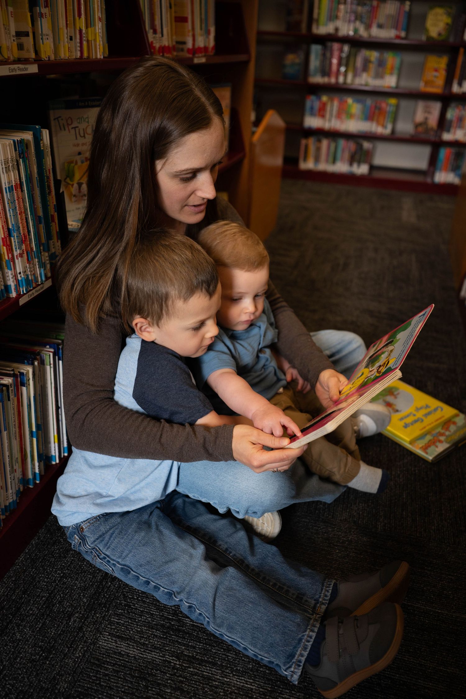 A mother reads a colorful picture book to her two young children while sitting on the floor of a library.