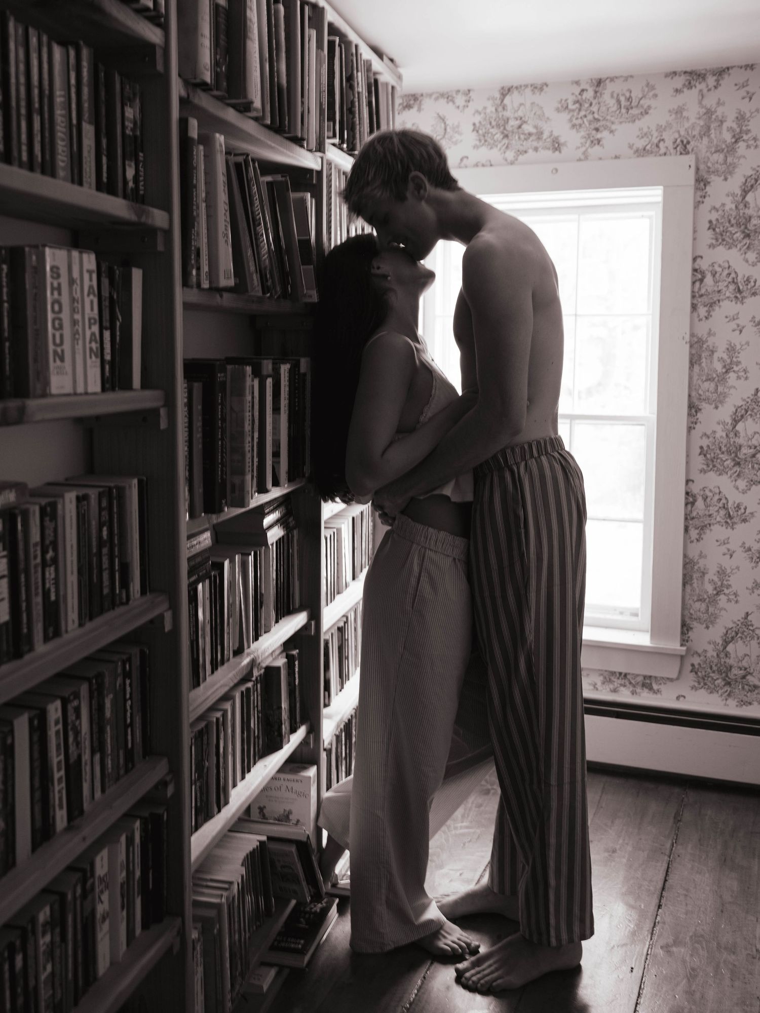 Black and white photo of couple sharing tender kiss beside tall bookshelf near bright window.
