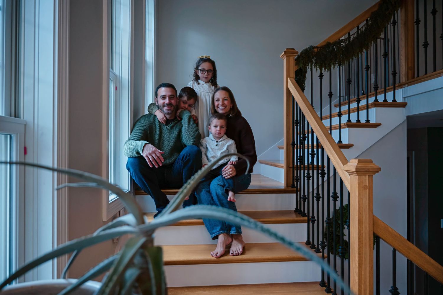 Family of five poses casually on home staircase with holiday garland and indoor plant in foreground.