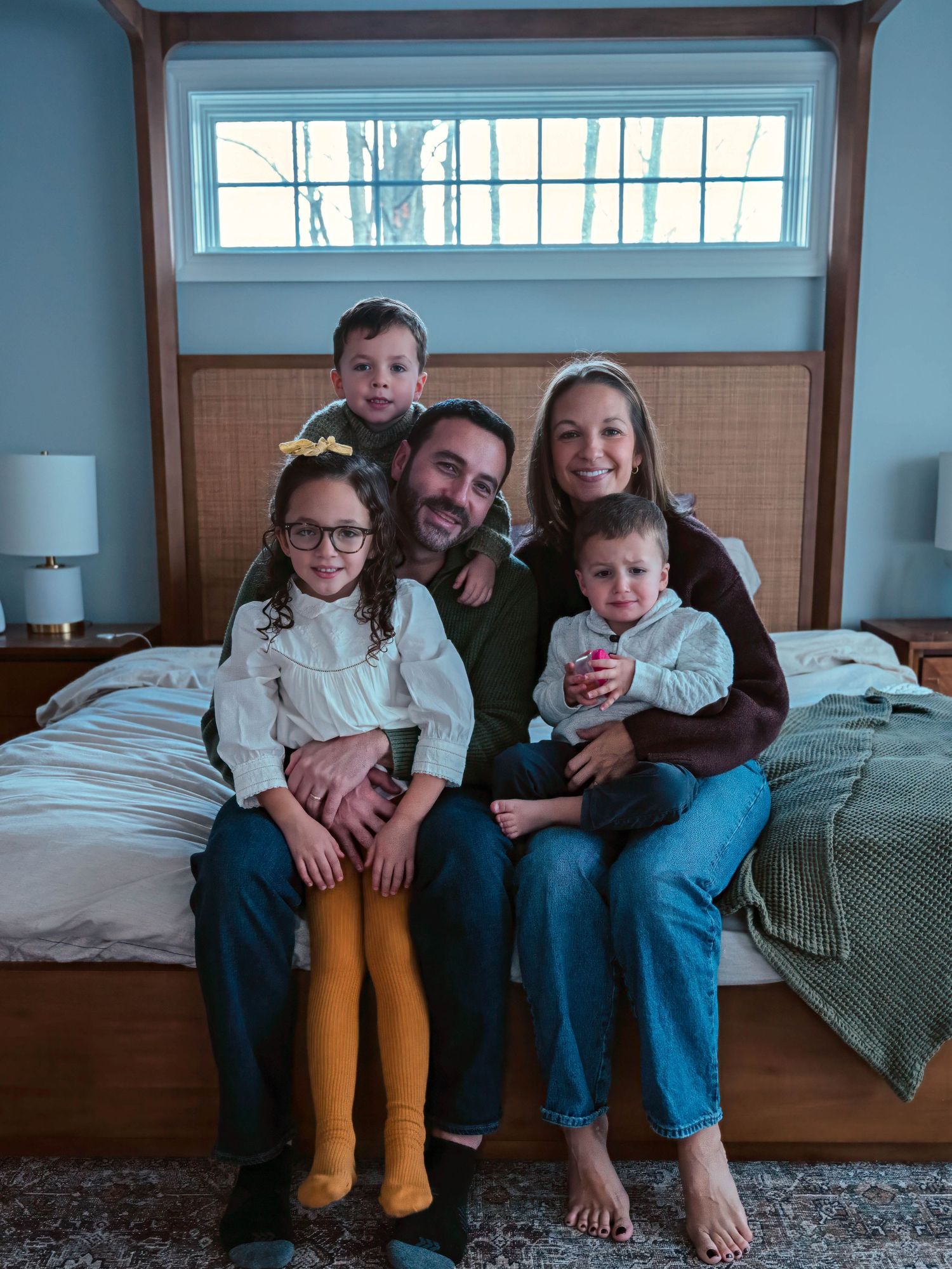 A happy family of five poses together on a bed in a cozy blue bedroom.