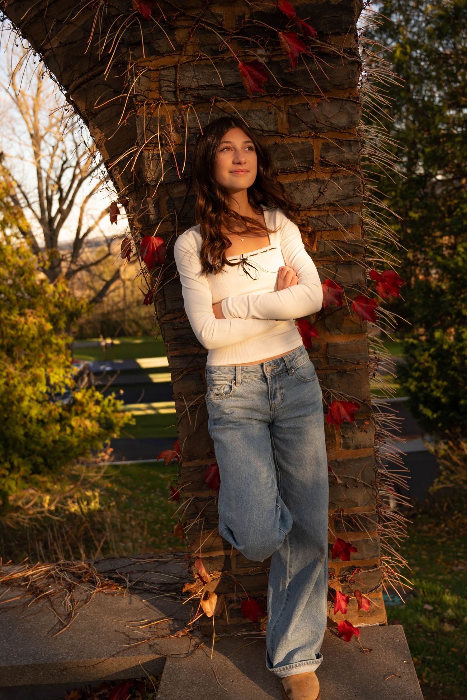 Teen girl in white crop top and jeans leaning against stone pillar with red autumn vines.