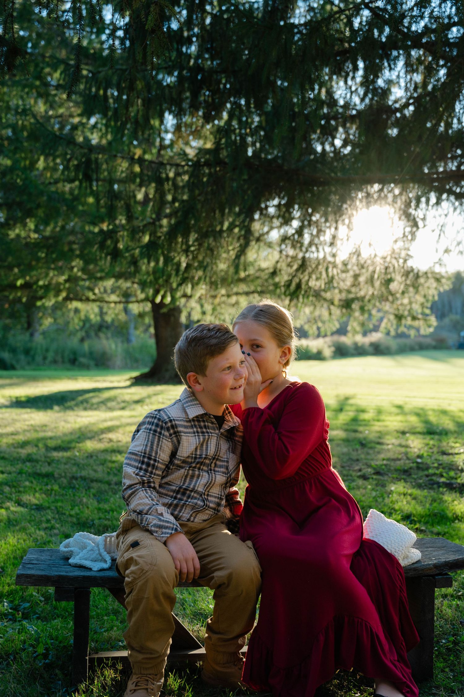 A boy and girl sit together on a wooden bench under a large tree in a sunlit green park, sharing a tender moment.
