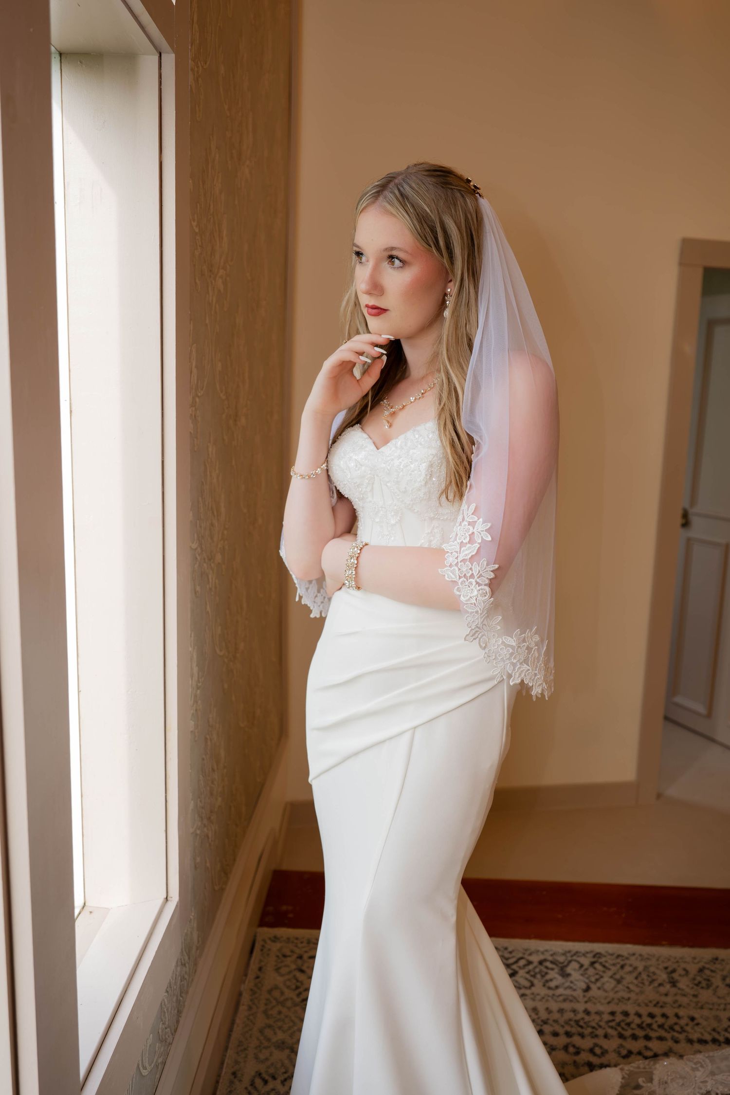 Elegant bride in a white mermaid gown and lace veil standing by a window in soft natural light.