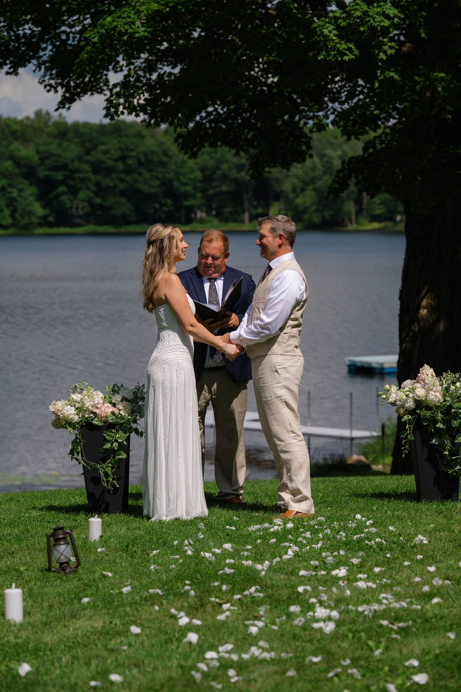 Outdoor lakeside wedding ceremony with bride in white gown, groom in tan suit, and officiant on green lawn.