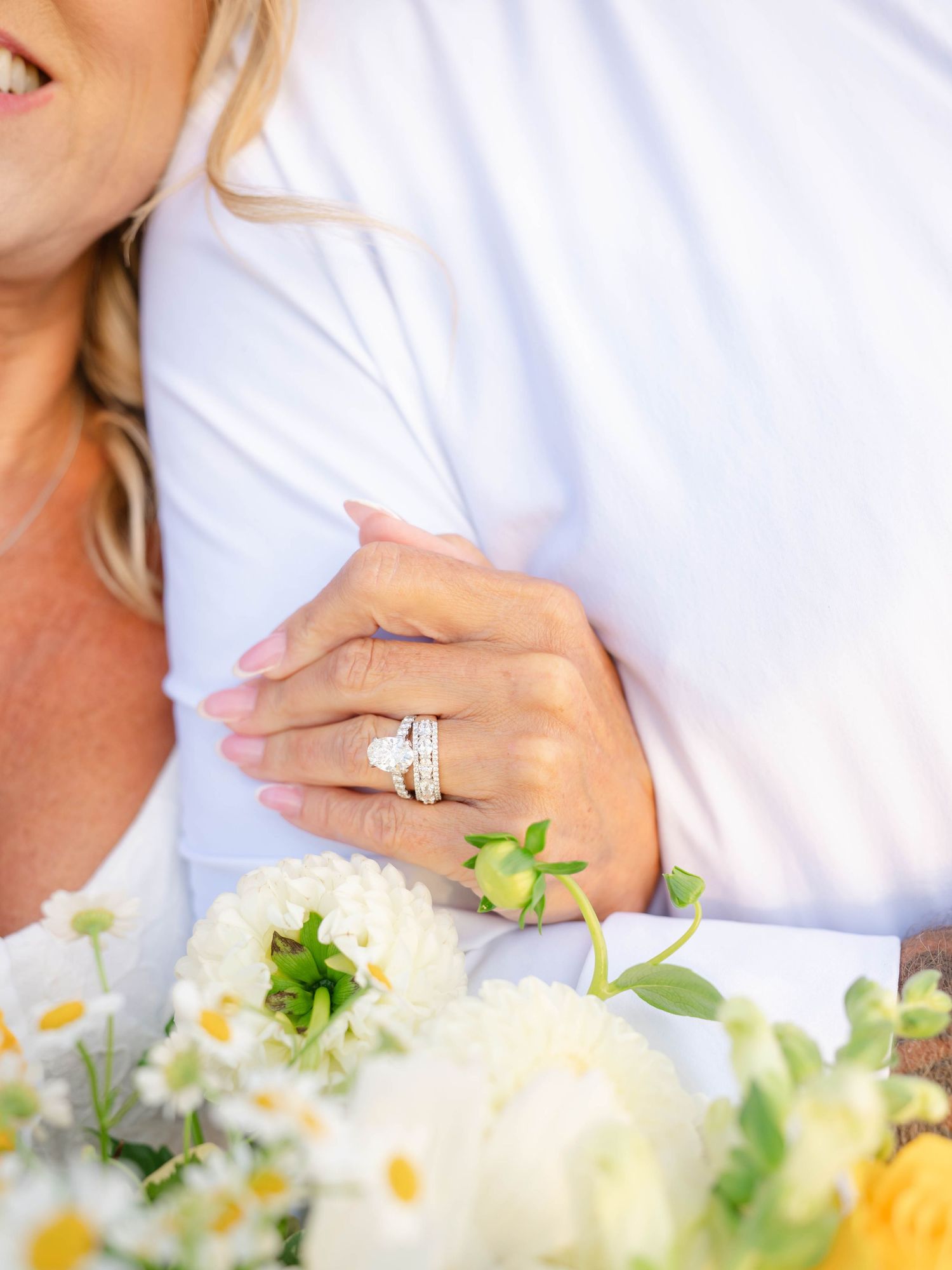 Close-up of a woman's hand displaying a diamond engagement ring and wedding band while holding white flowers.