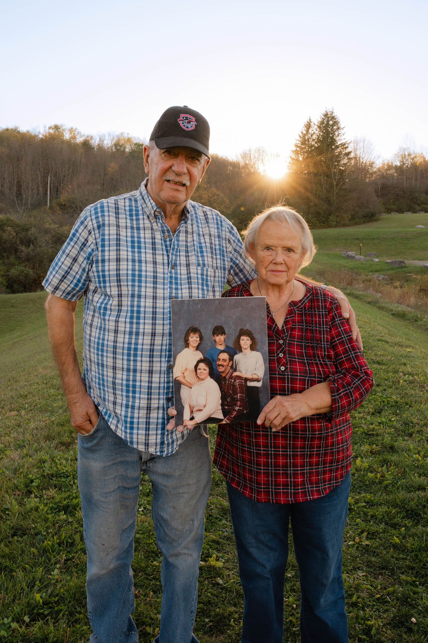 Senior couple holding old family photo outdoors at sunset with green hills behind them.