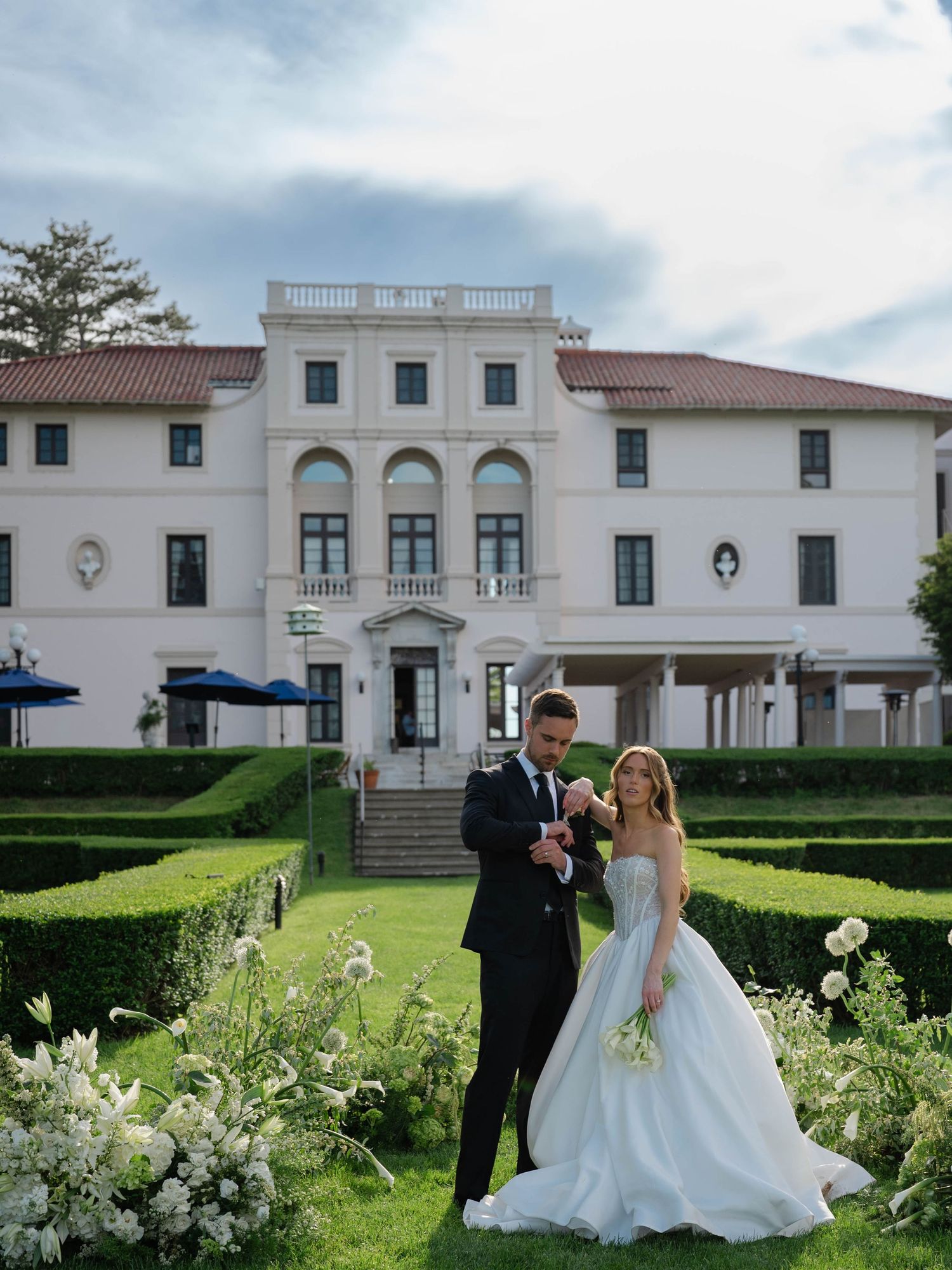 Bride in light blue ball gown and groom posing before grand white estate surrounded by formal gardens.