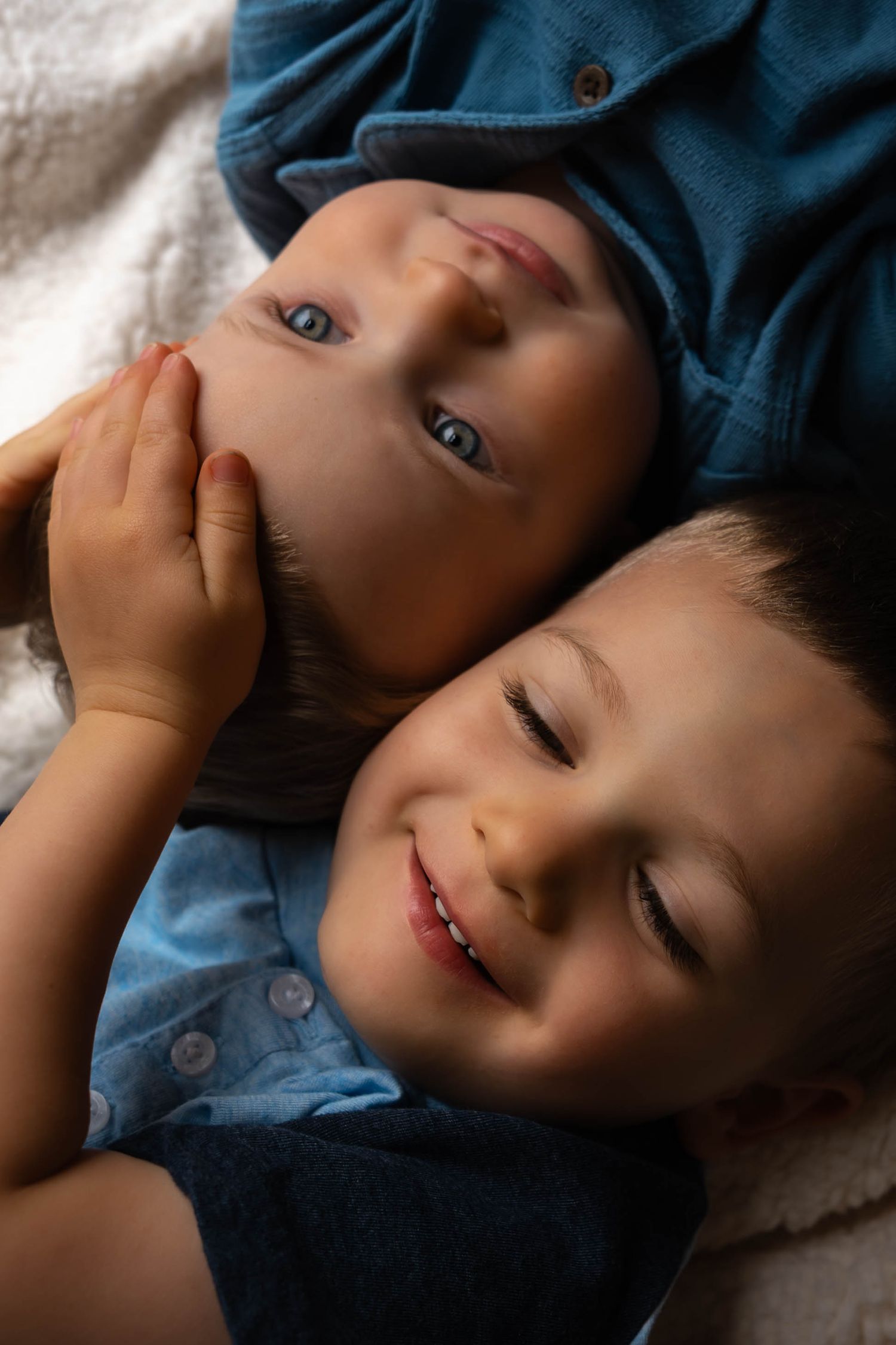 Close-up of two young brothers lying together, one smiling with eyes closed, on a blanket.
