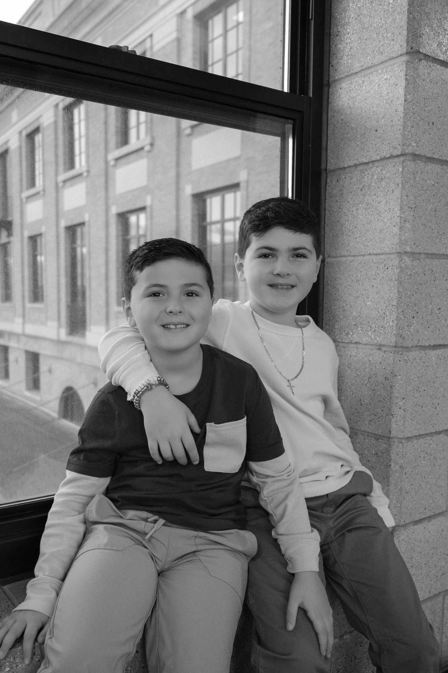 Black and white portrait of two young brothers sitting together smiling by a large window outdoors.