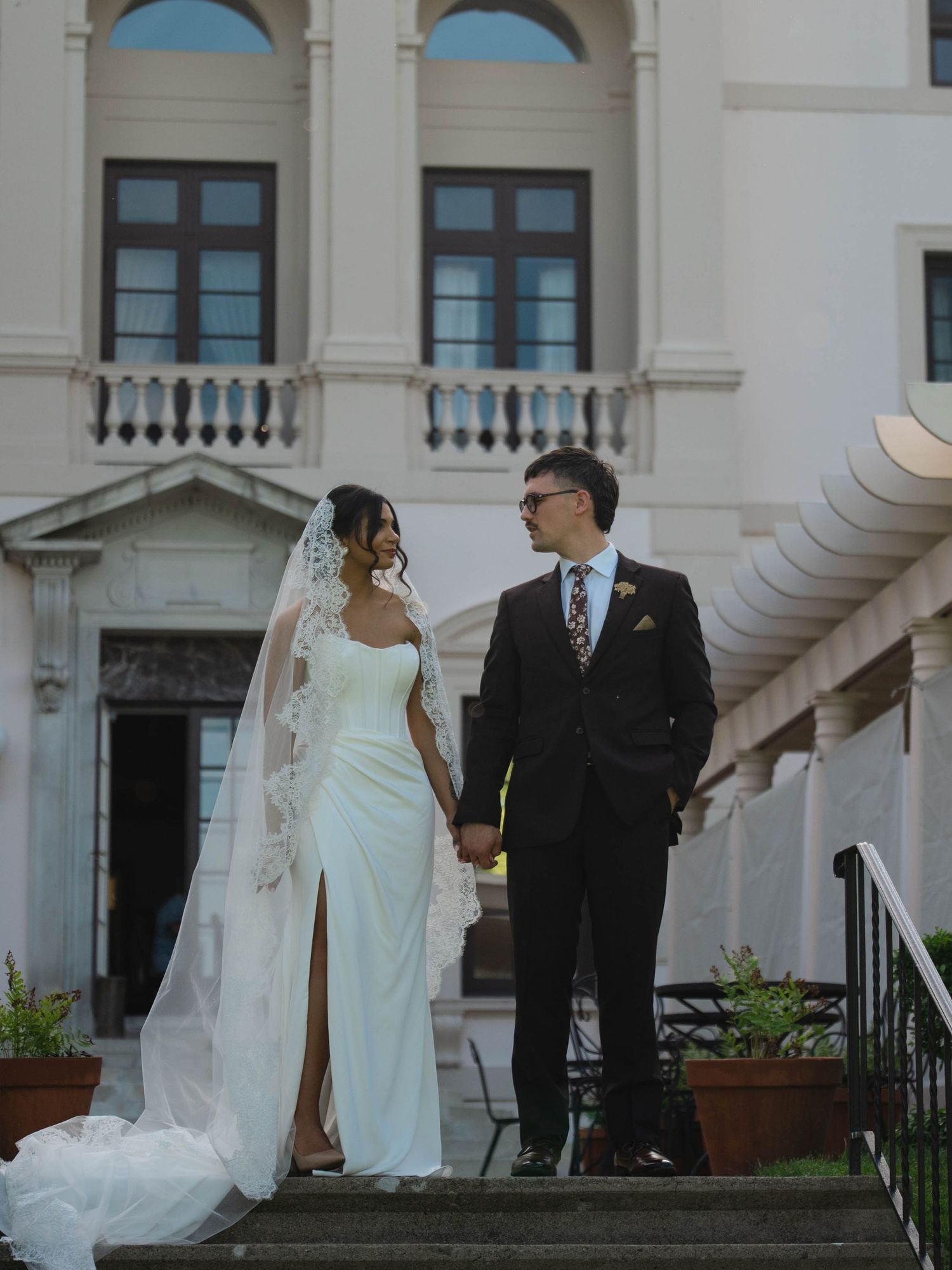 Bride and groom holding hands on steps of grand classical building with arched windows and balcony.