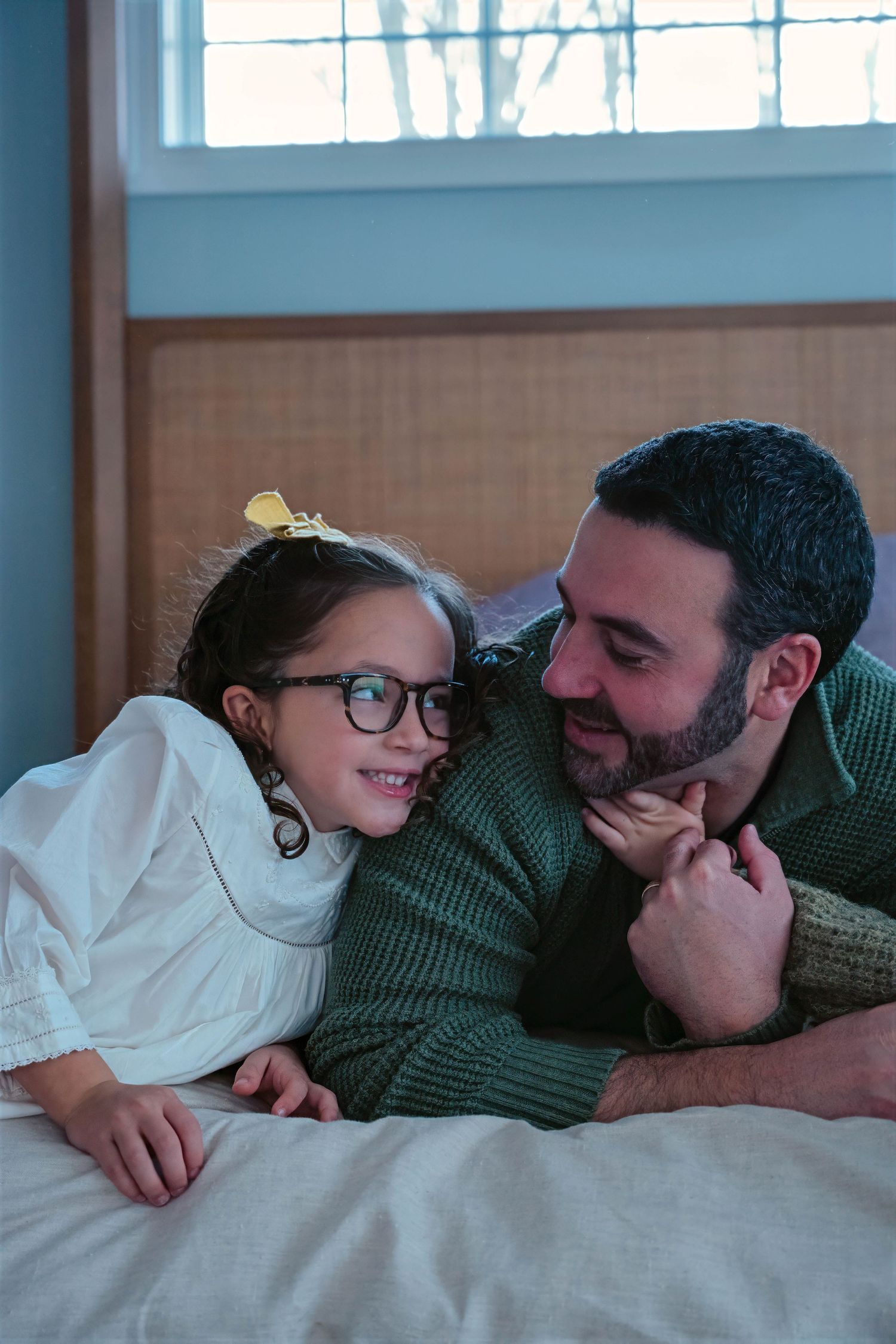 A smiling girl with glasses and a yellow bow shares a joyful moment with her dad on a bed.