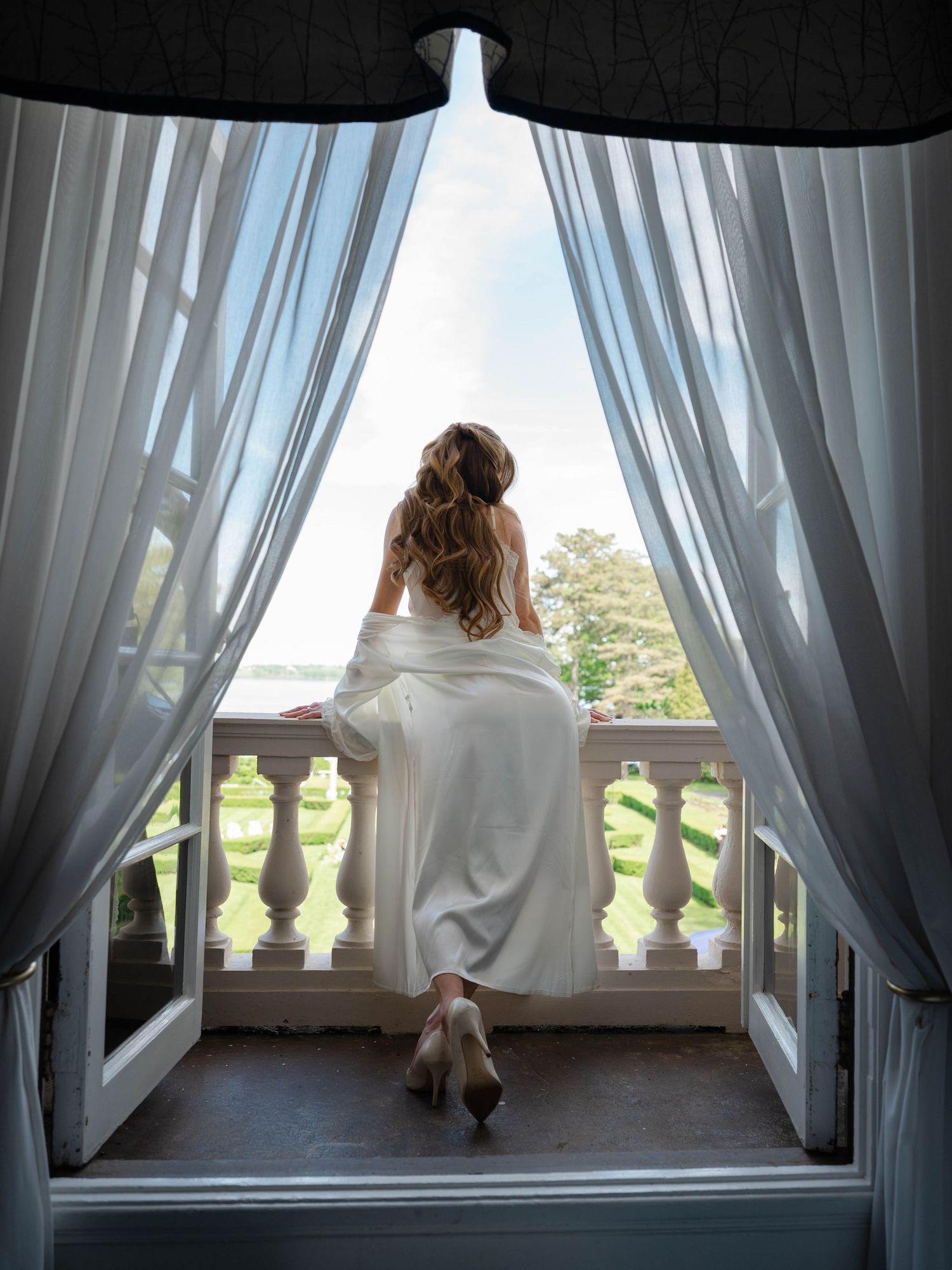 Bride in white gown standing on an ornate balcony framed by flowing curtains overlooking a garden.