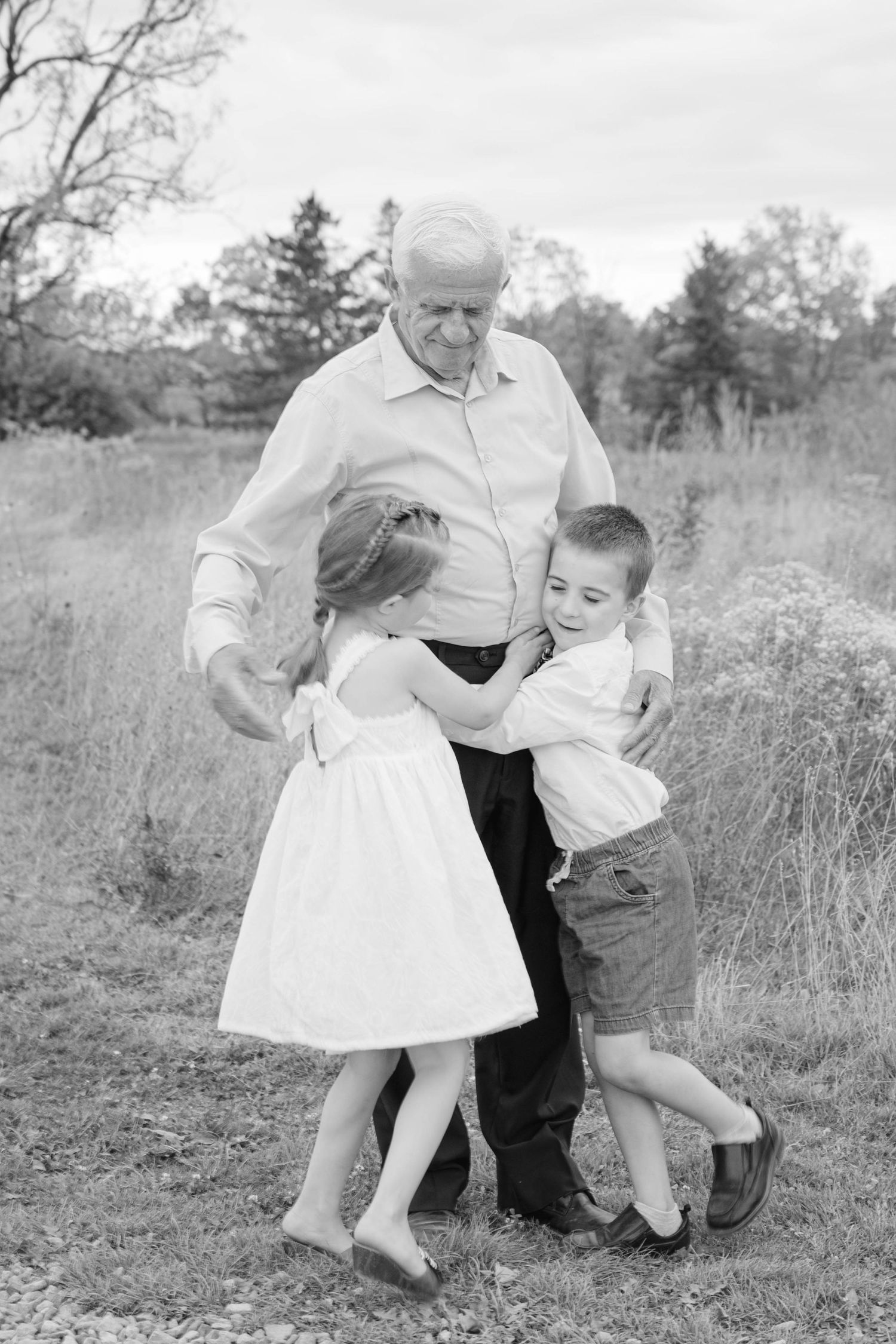 Black and white photo of two young children hugging their grandfather outdoors in a natural field setting.