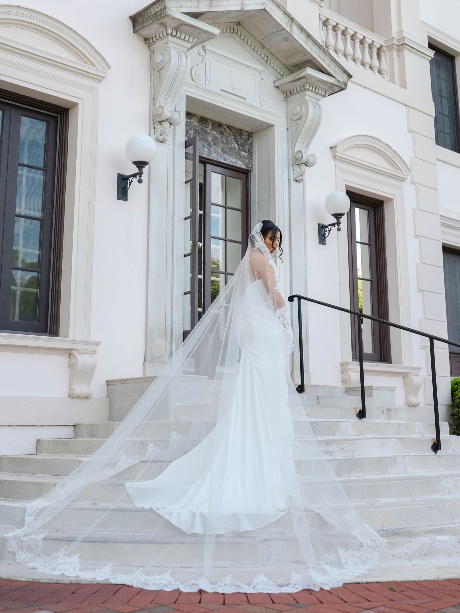 Bride in fitted mermaid wedding dress with dramatic lace-edged cathedral veil posing on steps of a white neoclassical building. Finger lakes