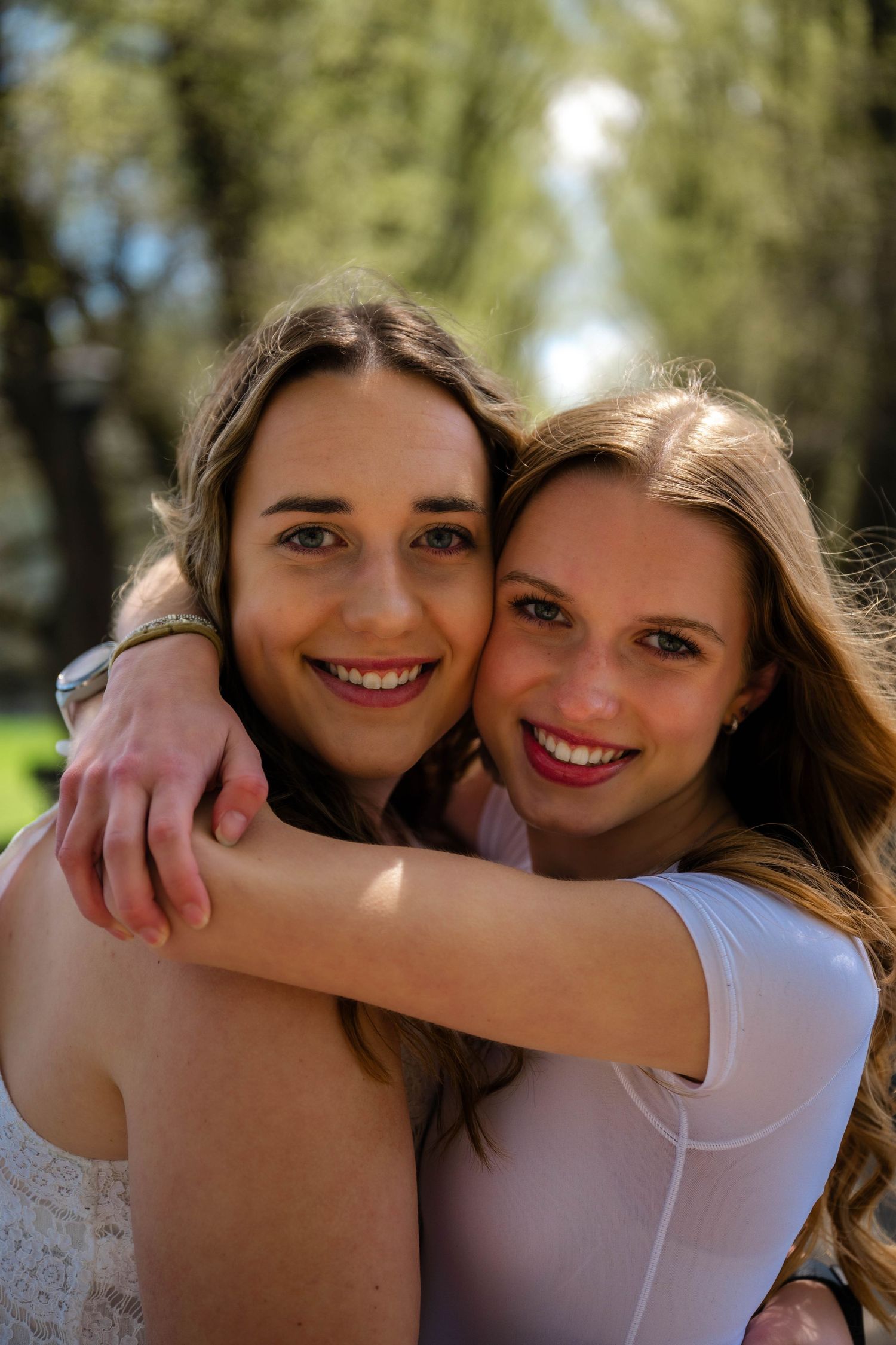 Two smiling young women with brown hair embrace warmly outdoors in a sunny park setting.