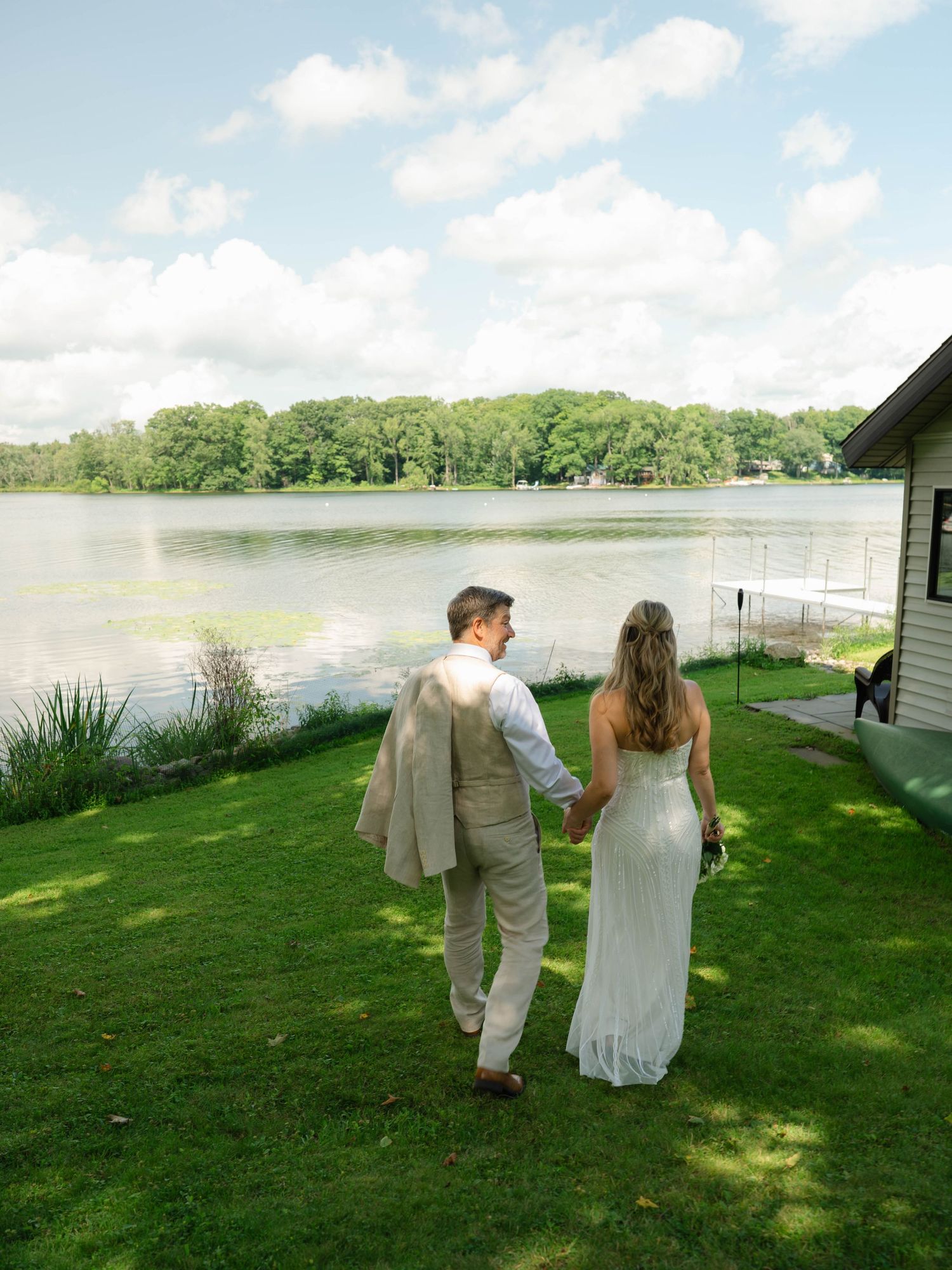 Bride and groom walk hand-in-hand toward a serene lakeside on lush green grass on their wedding day.