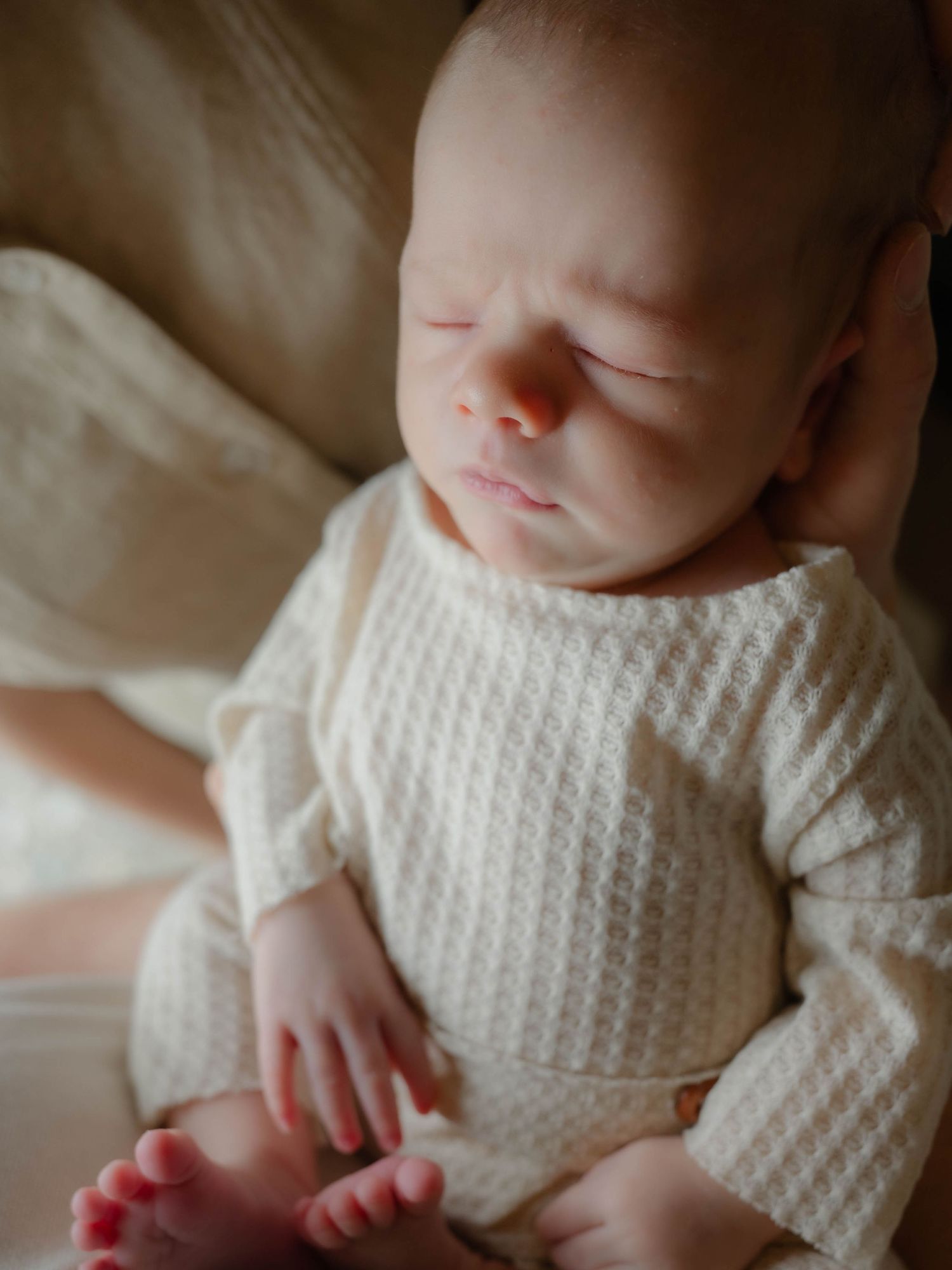 Peaceful newborn baby sleeping in cream waffle knit onesie, tiny fingers visible, resting on a soft beige cushion.