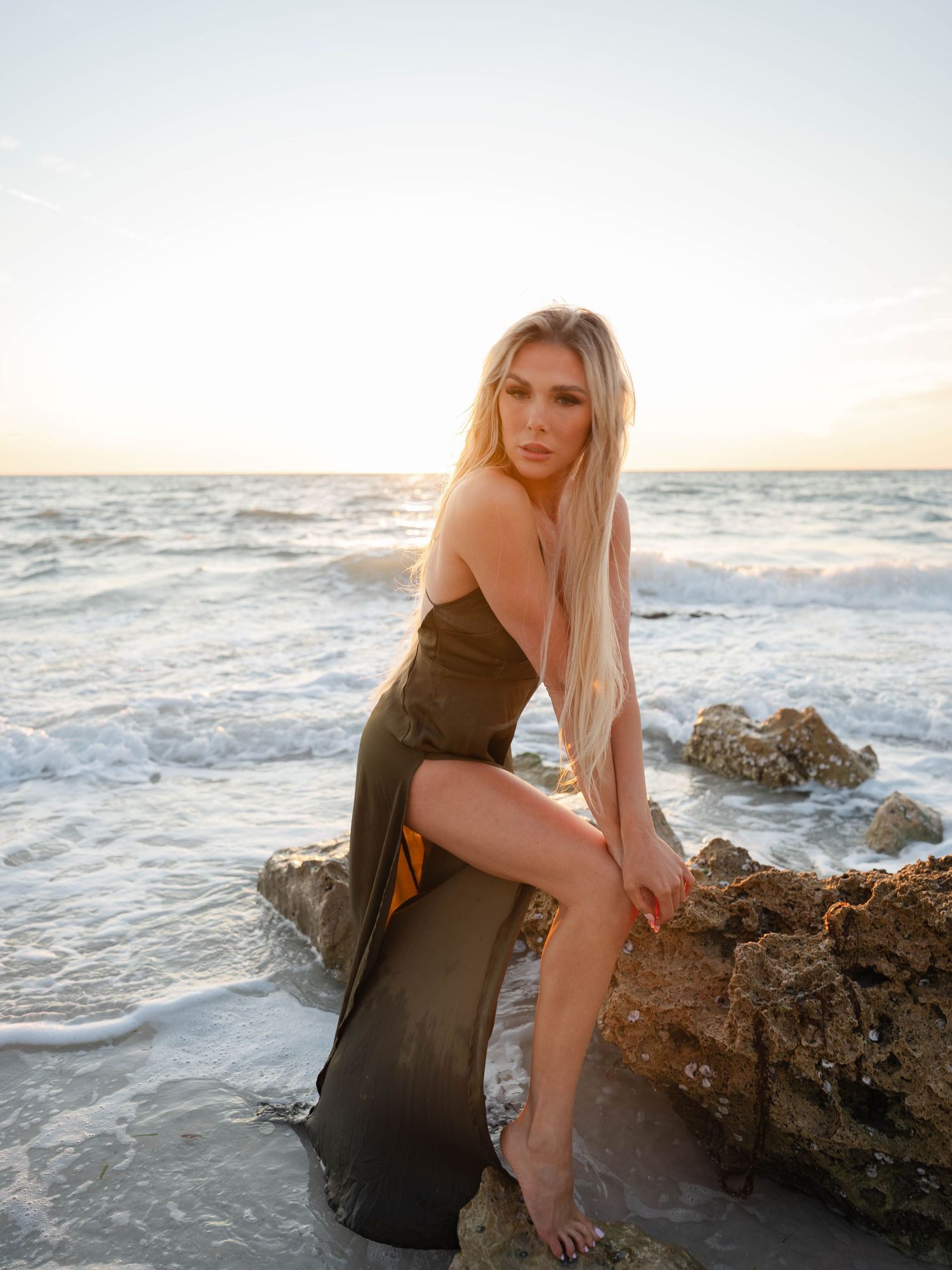 Blonde woman in olive satin dress sitting on beach rocks at golden sunset.