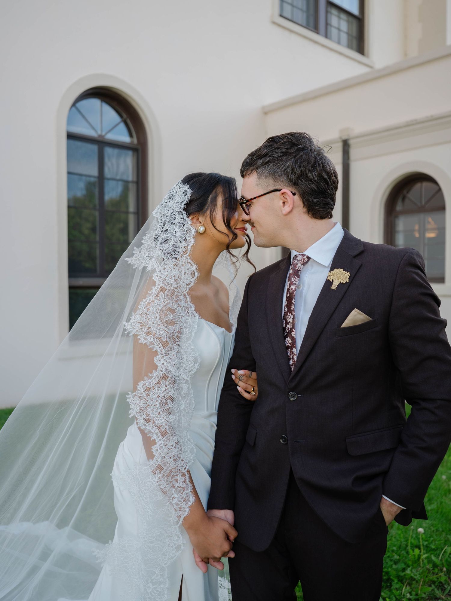 Bride and groom share a kiss outside white estate, bride wearing long lace-trimmed cathedral veil.