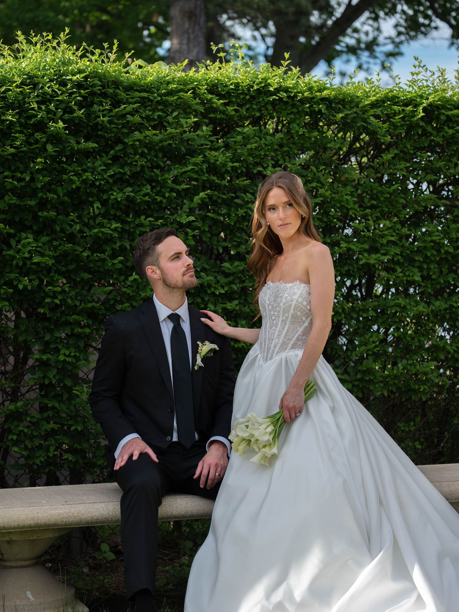 Bride in strapless lace gown holds calla lilies beside seated groom against a lush green hedge backdrop.