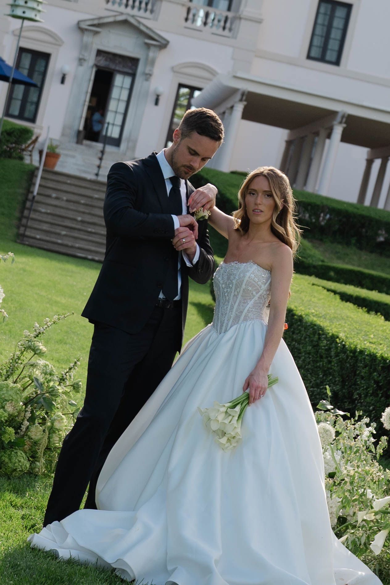 Groom adjusting bride's jewelry as she holds calla lily bouquet in garden before white manor house.