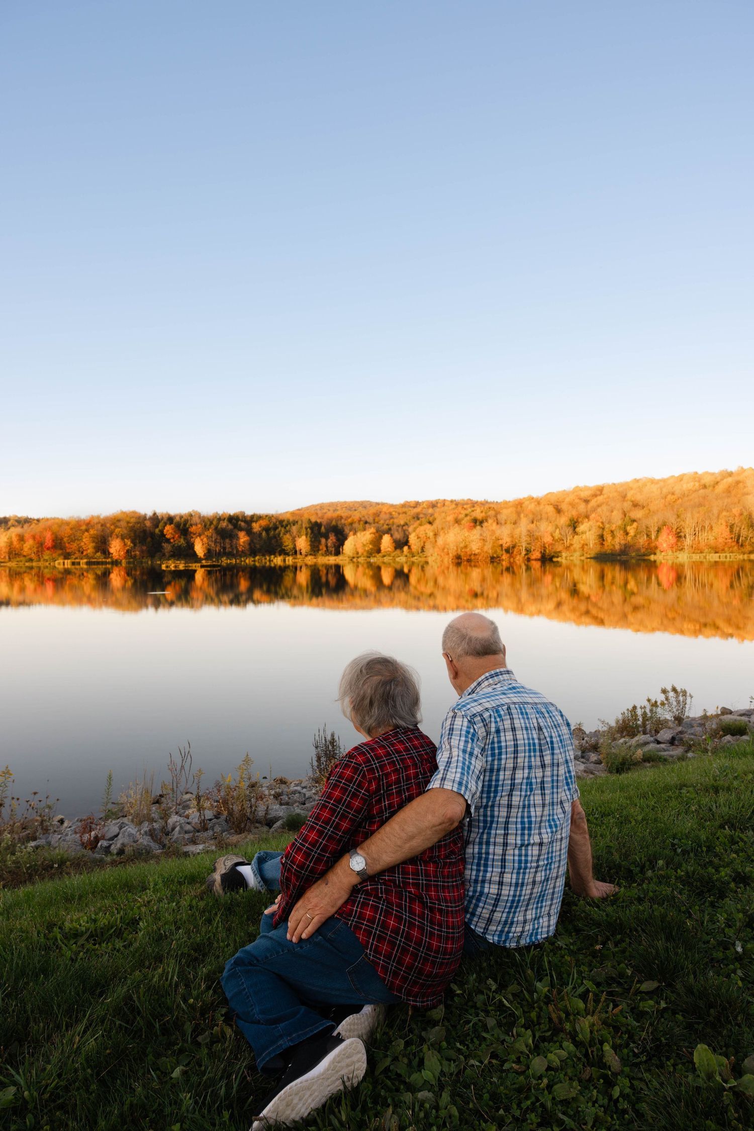Elderly couple sitting together arm in arm by a serene lake with colorful fall foliage reflection.