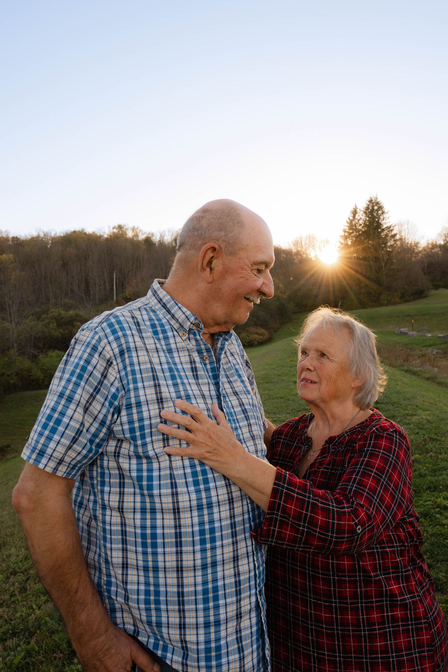 Elderly couple smiling at each other outdoors at sunset on a green hillside in plaid shirts.