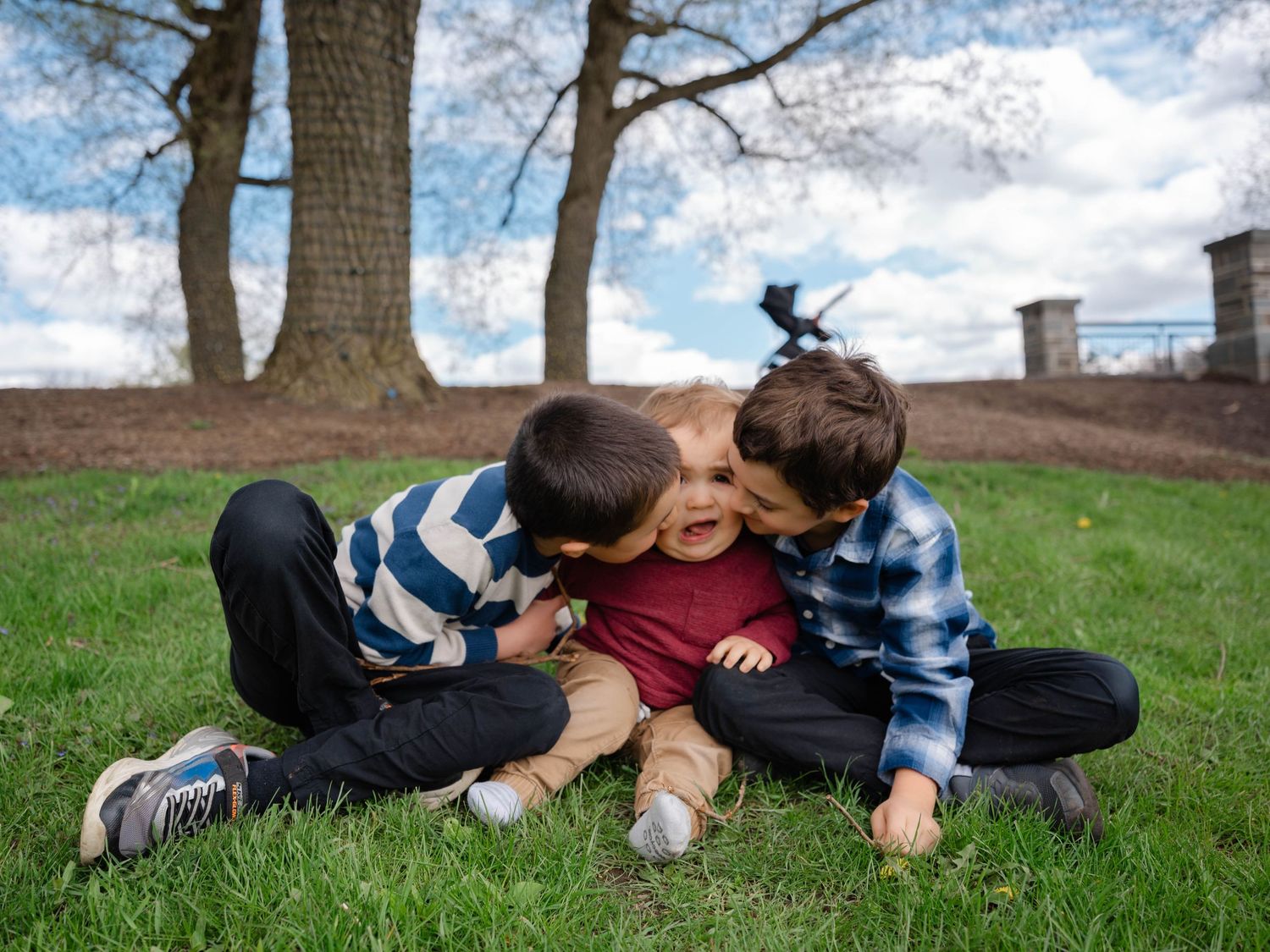 Two older brothers giving kisses to baby sibling while sitting on grass in a park.