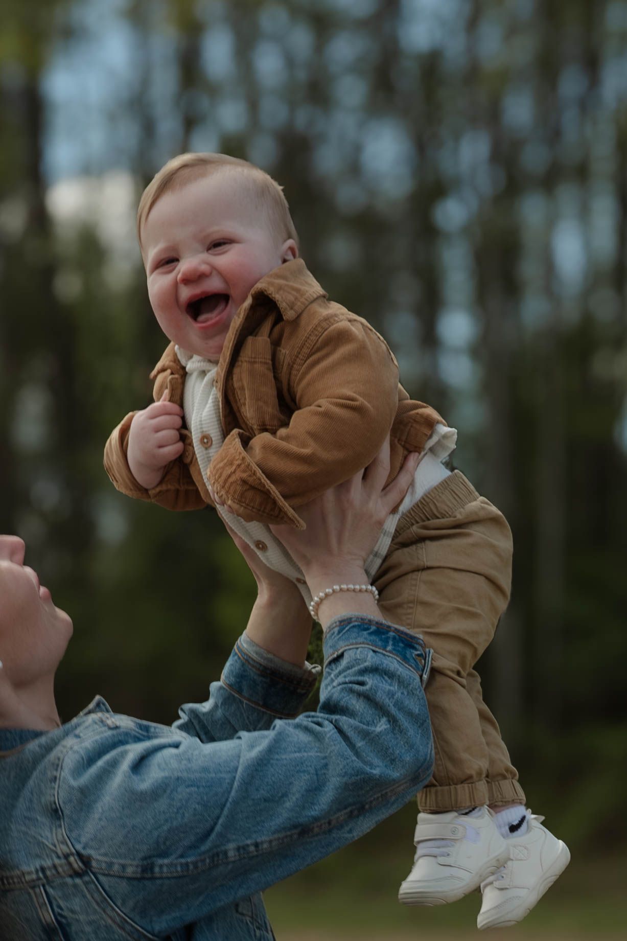 Laughing baby in brown outfit being lifted up outdoors by parent in denim.