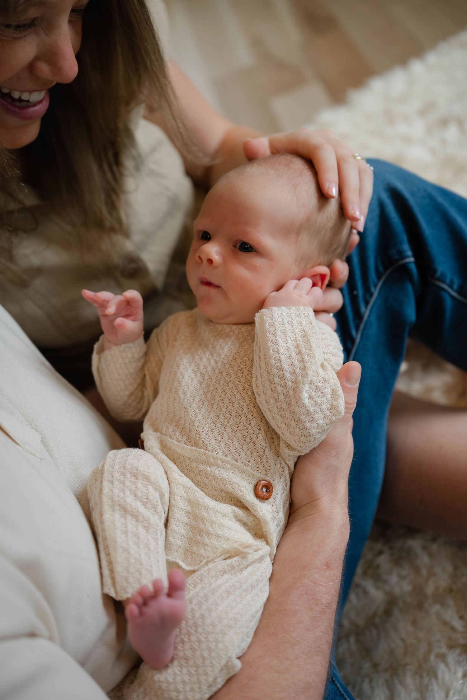 Parents lovingly cradling their newborn baby dressed in a cream knit romper on a cozy carpet floor.