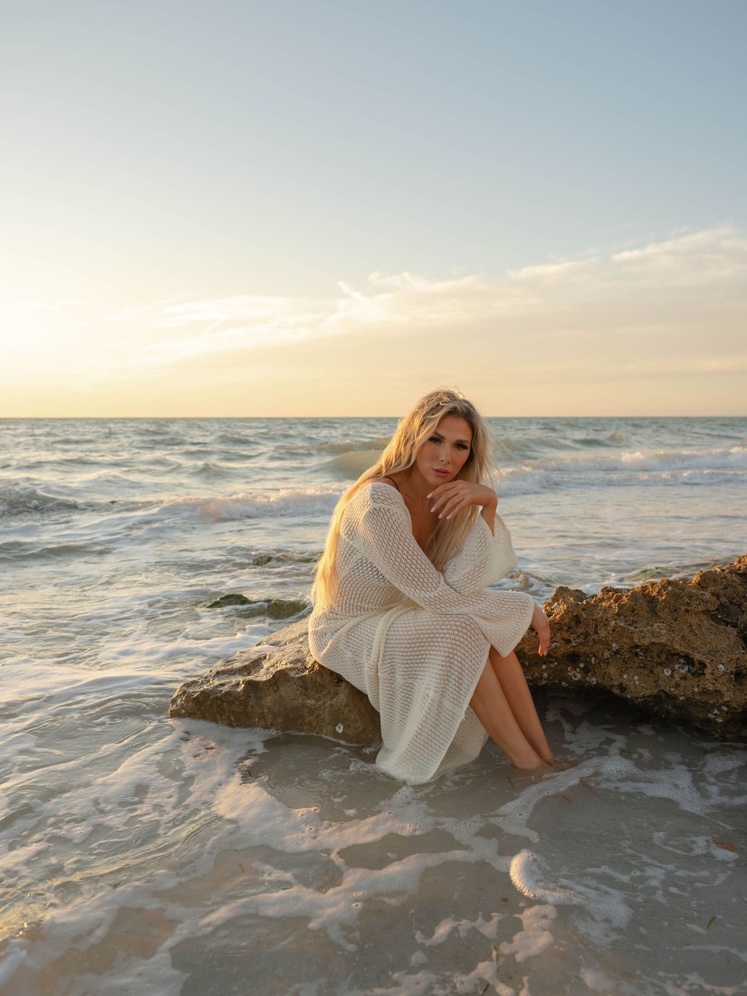 Blonde woman in white knit dress sitting on ocean rocks during sunset.