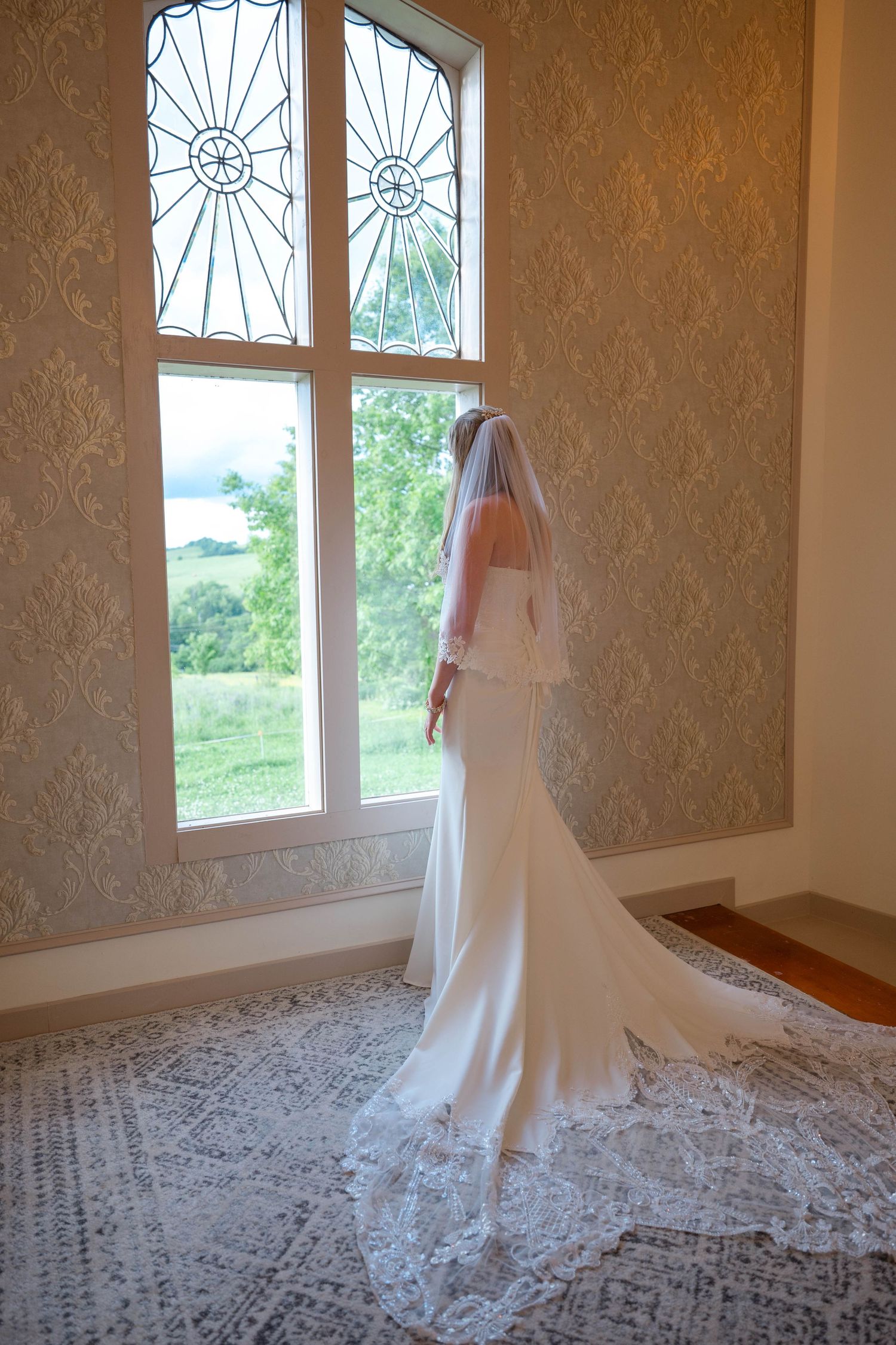 Bride in white gown and veil stands before ornate stained glass window, with lace train spread across tiled floor.