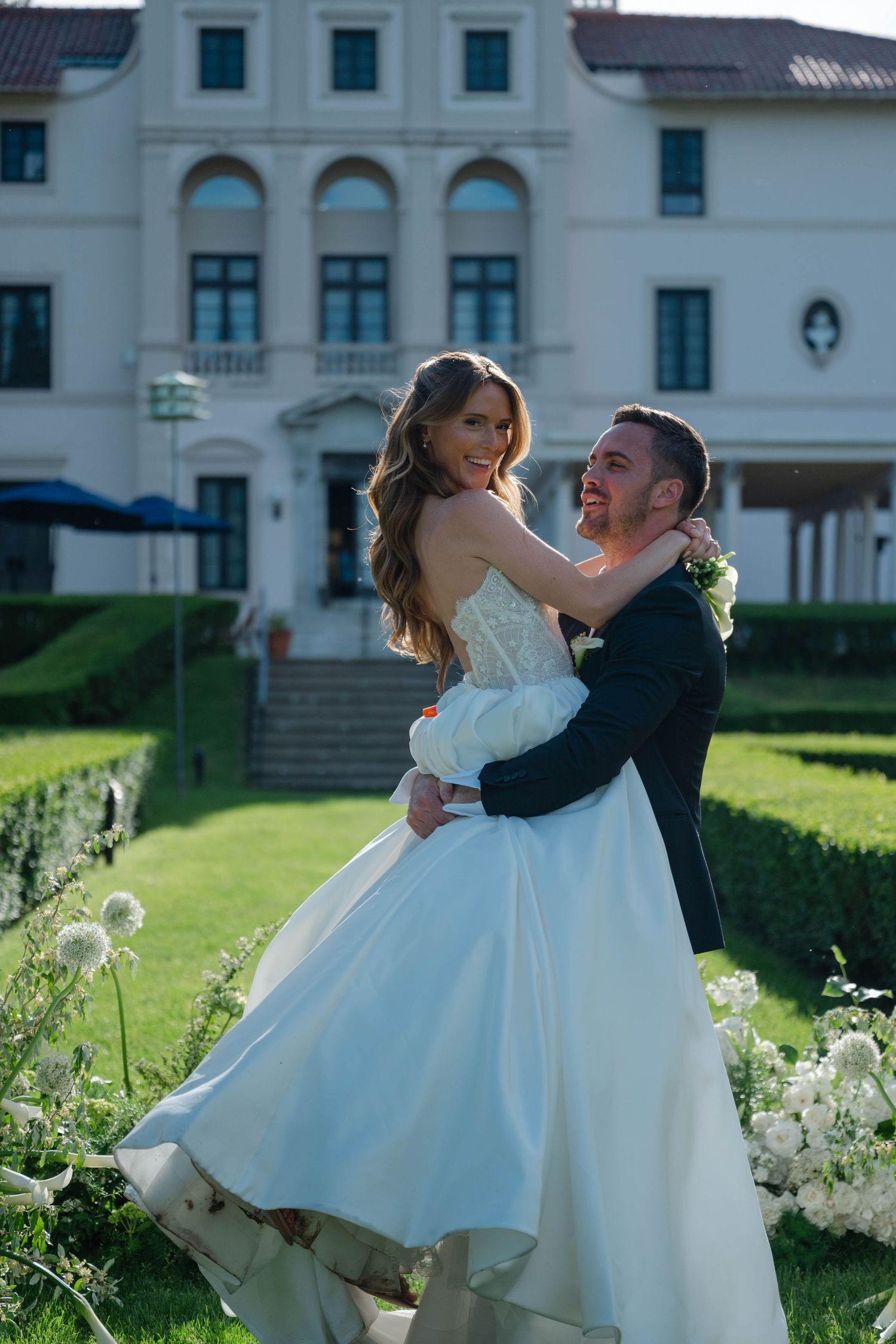 Groom lifting smiling bride in strapless white gown in manicured garden with white estate backdrop.