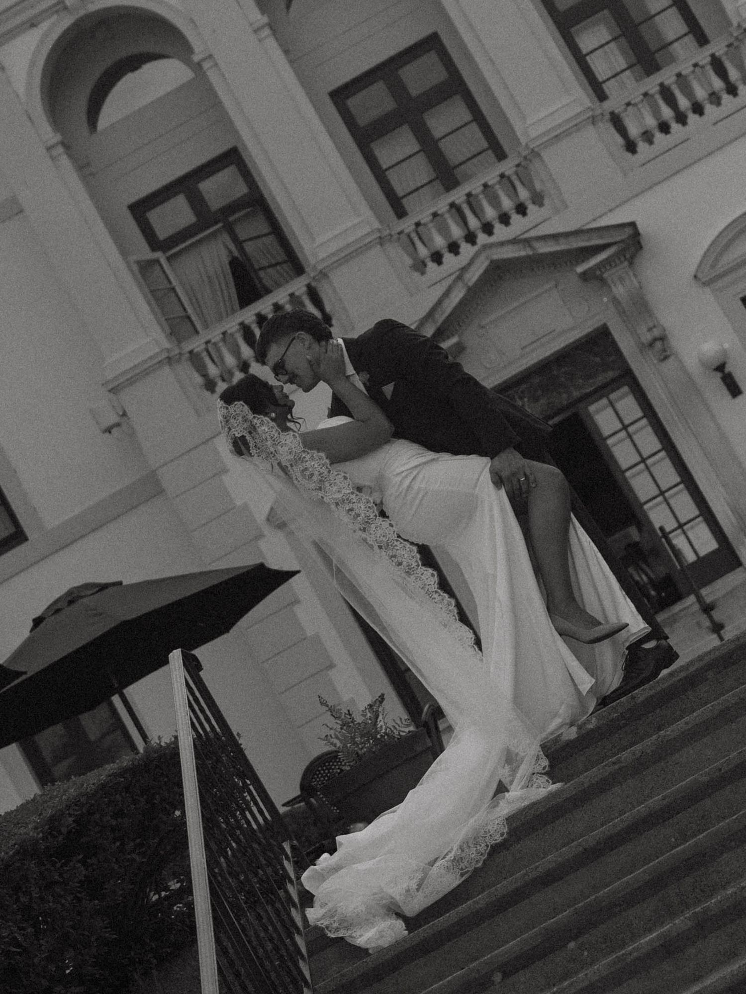 Black and white photo of groom dipping bride in wedding dress on grand staircase steps.