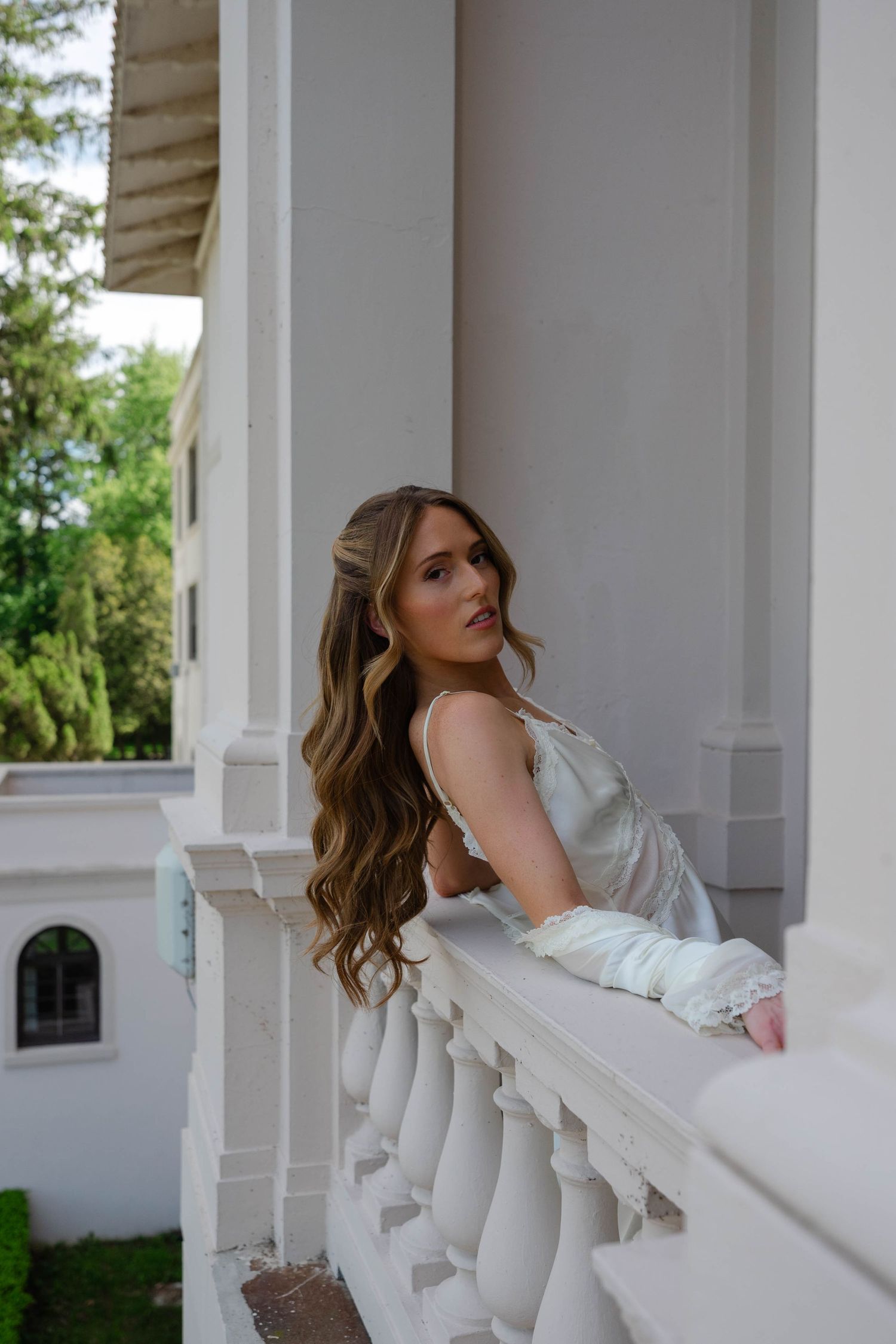 Elegant bride in white slip dress with long wavy hair leans on white balcony railing outdoors.