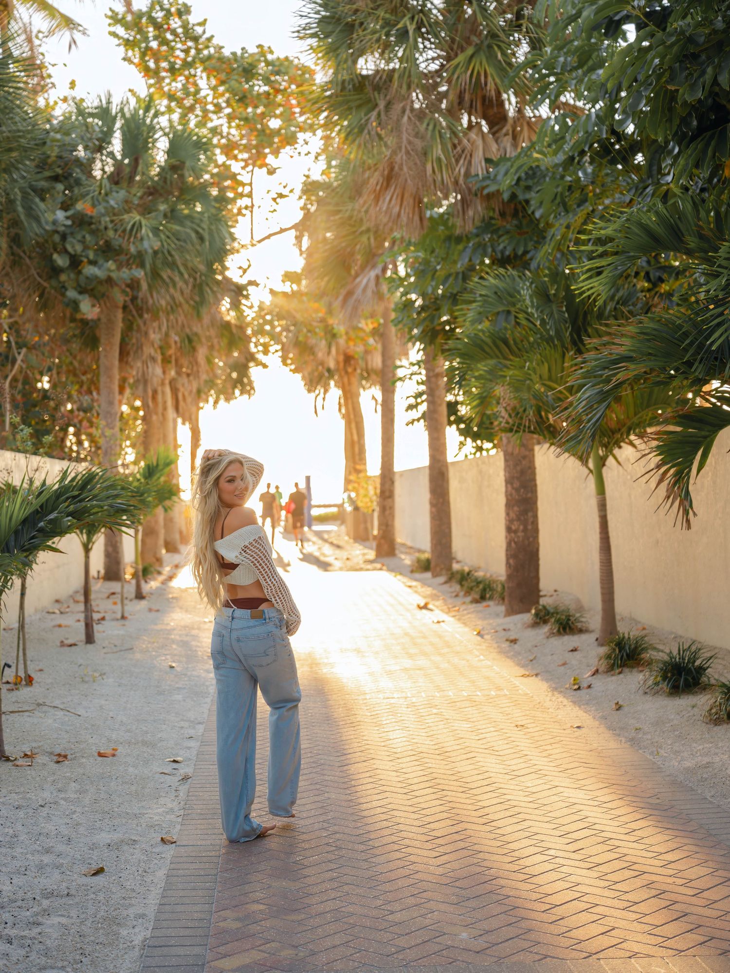 Blonde woman in jeans and white top walking along a sunlit palm-lined brick pathway toward the beach.