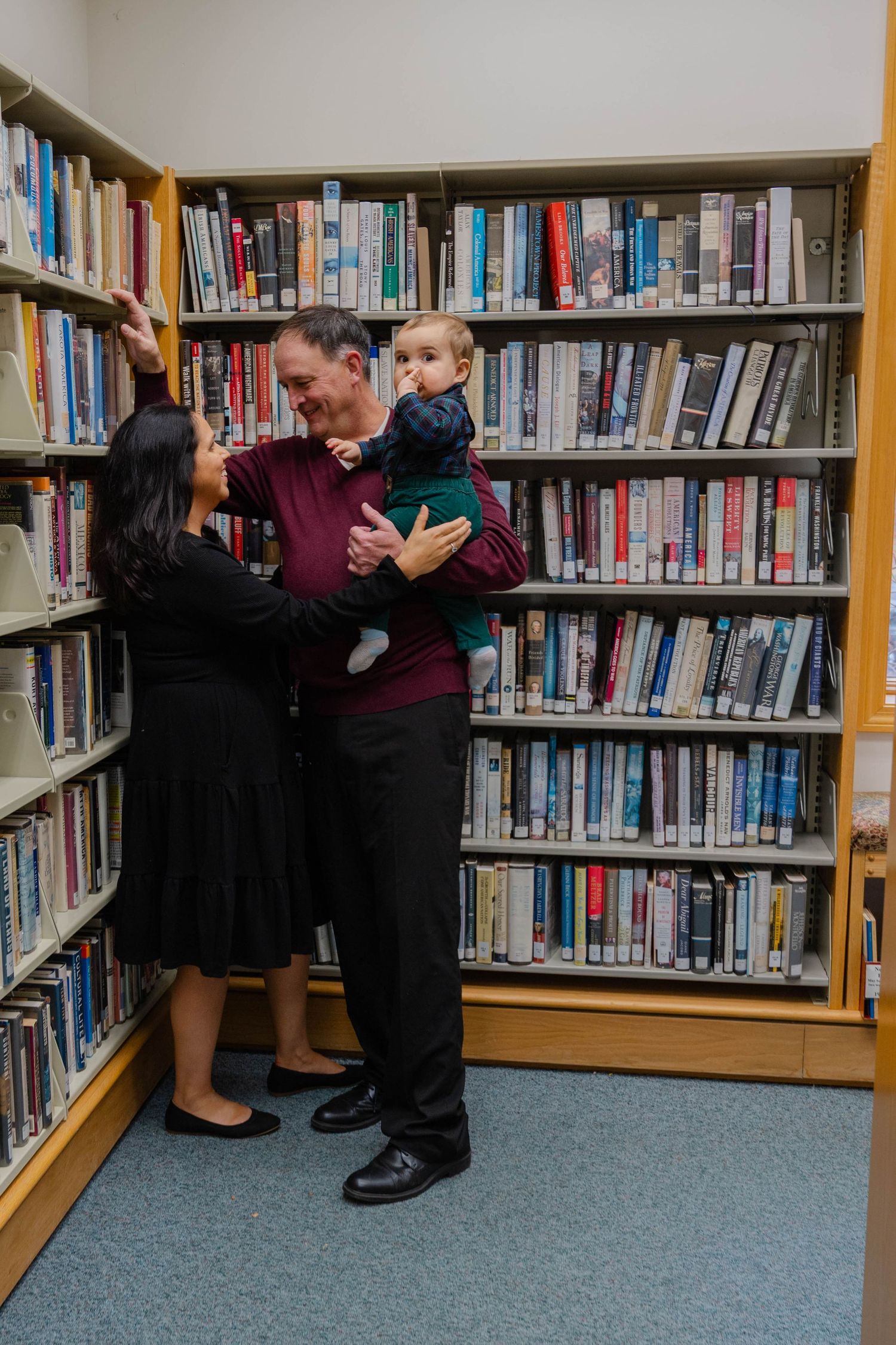 Smiling couple with baby boy standing near bookshelves inside a cozy library setting.
