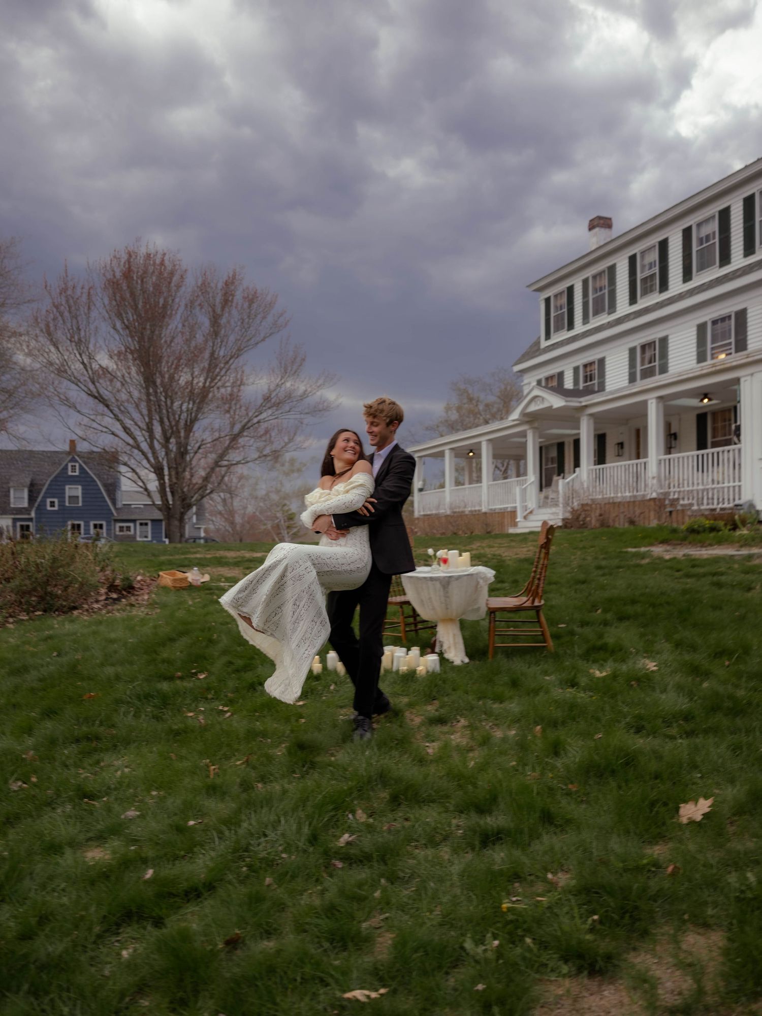 Groom lifting bride in white dress outdoors at elegant estate wedding venue under dramatic cloudy sky. Maine Wedding