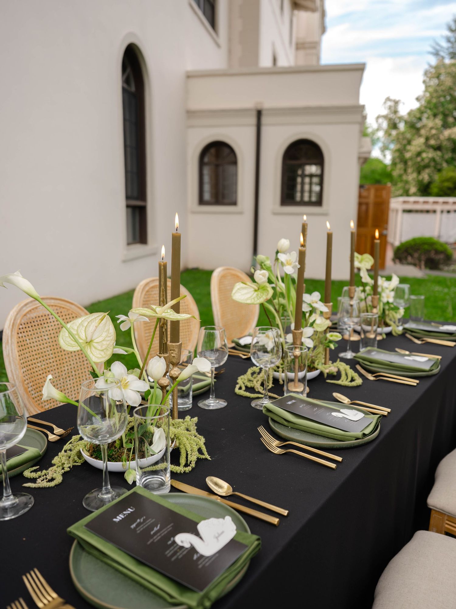 Elegant outdoor wedding reception table with black linen, gold cutlery, green napkins, white florals, and tall taper candles.