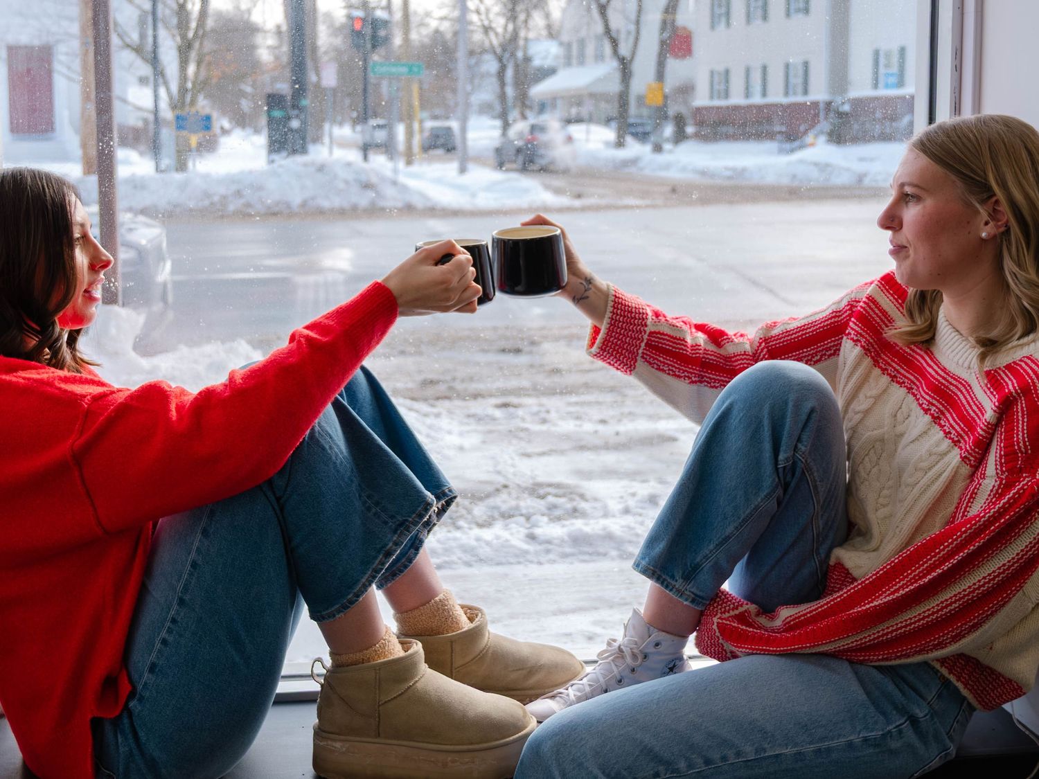 Two women toasting mugs of coffee by a snowy winter window in a cozy cafe.