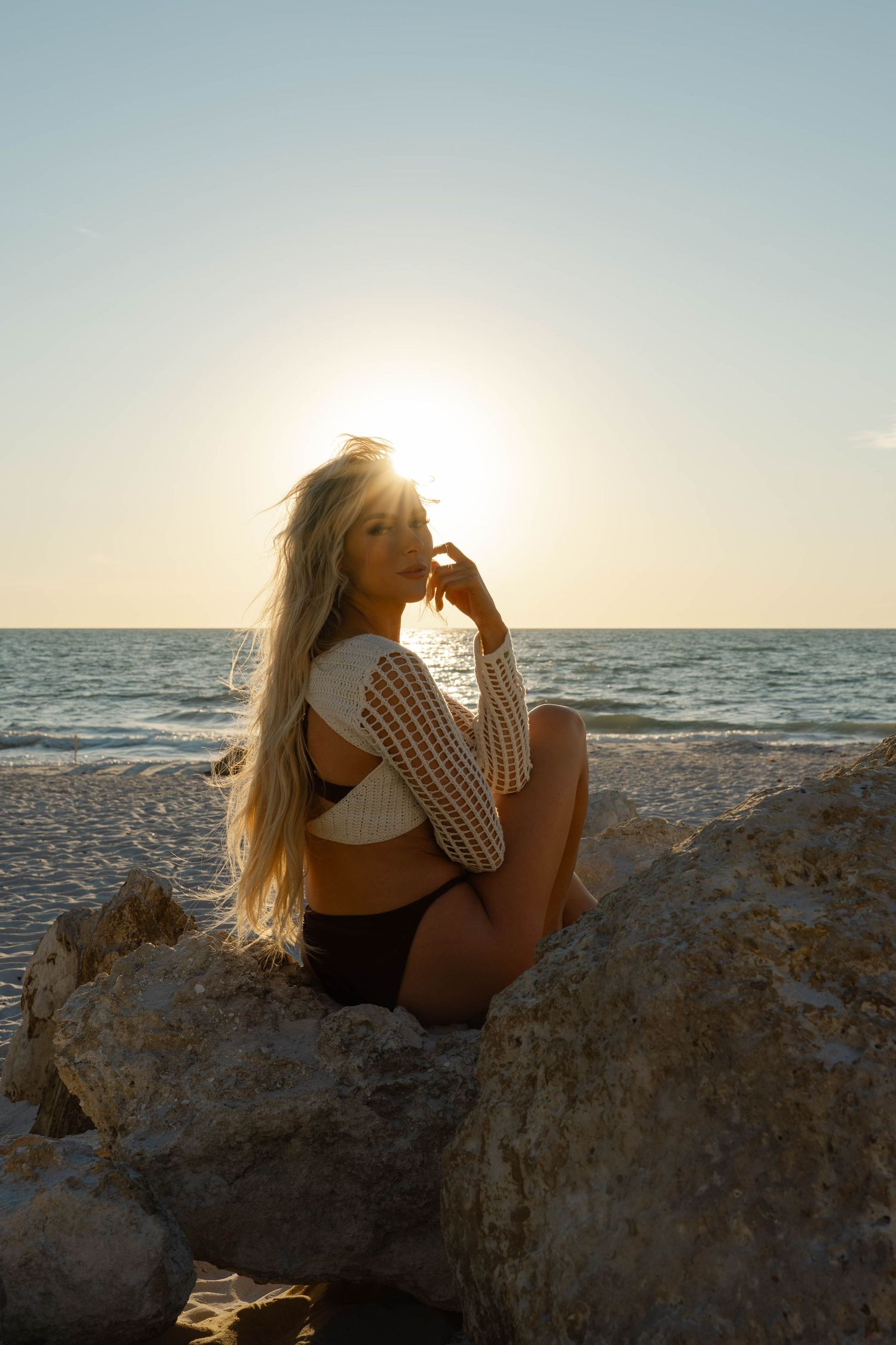 Blonde woman in bikini and white mesh top sitting on rocks at the beach with the sun setting over the ocean.