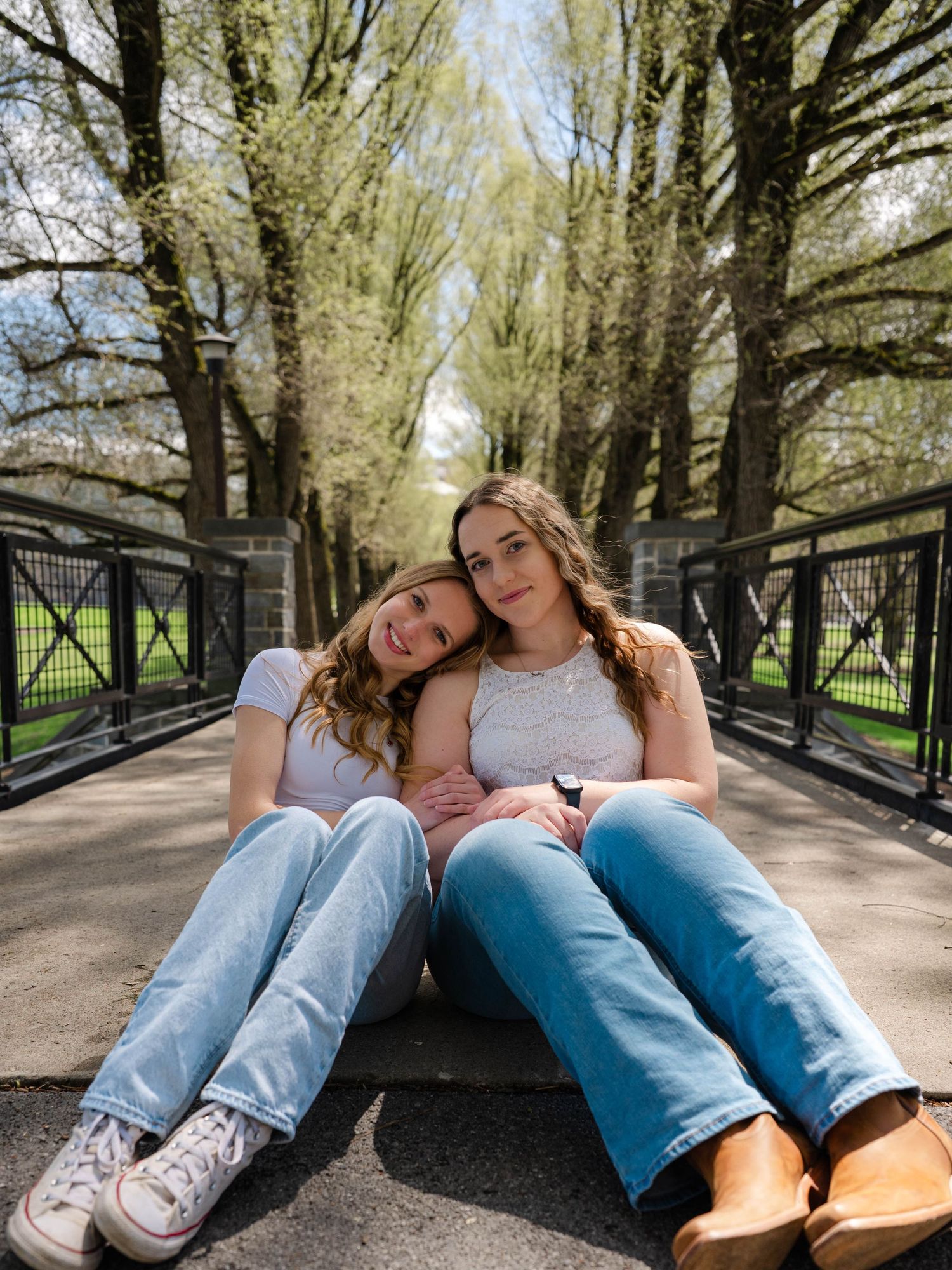 Two young women sit closely together on a bridge path surrounded by tall trees in a park.