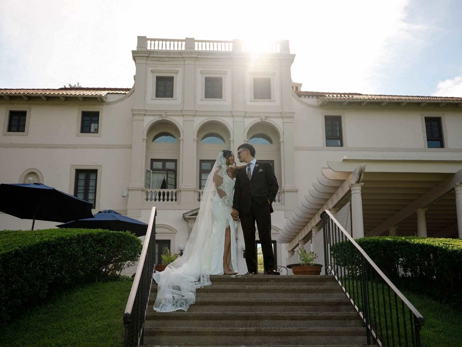 Bride and groom stand on sunlit steps of stunning white wedding venue with manicured hedges. Finger Lakes Region