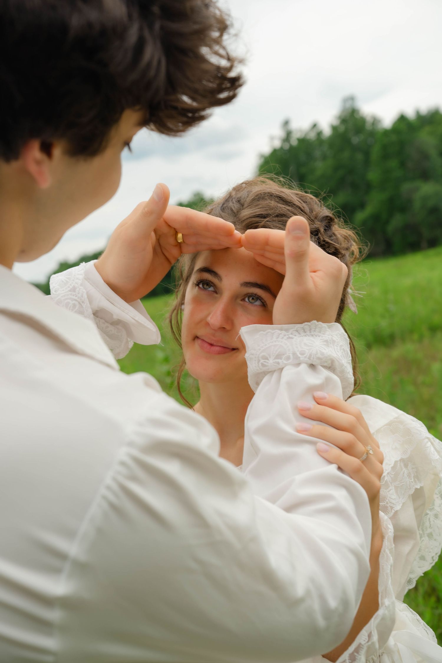 Romantic couple in white outfits framing face with hands in lush green field, wedding portrait photography.