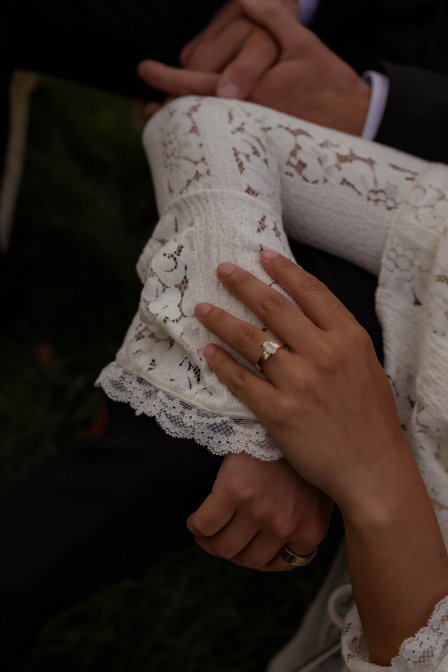 Bride wearing a diamond ring and lace wedding dress sleeve, with groom in suit visible in background. Maine Wedding Couple photography