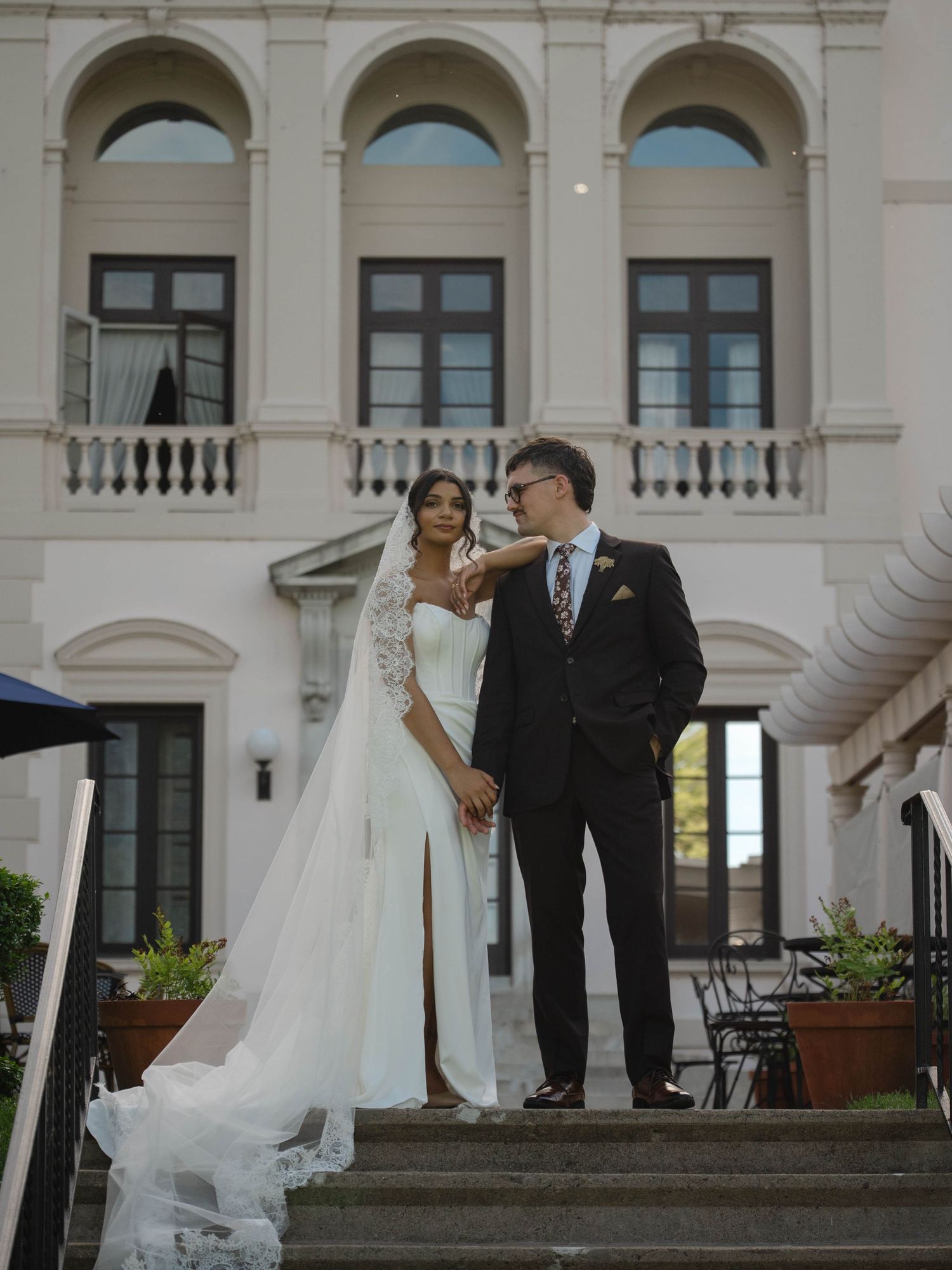 Elegant wedding couple poses on steps before grand white Italian-style mansion, bride in flowing veil.