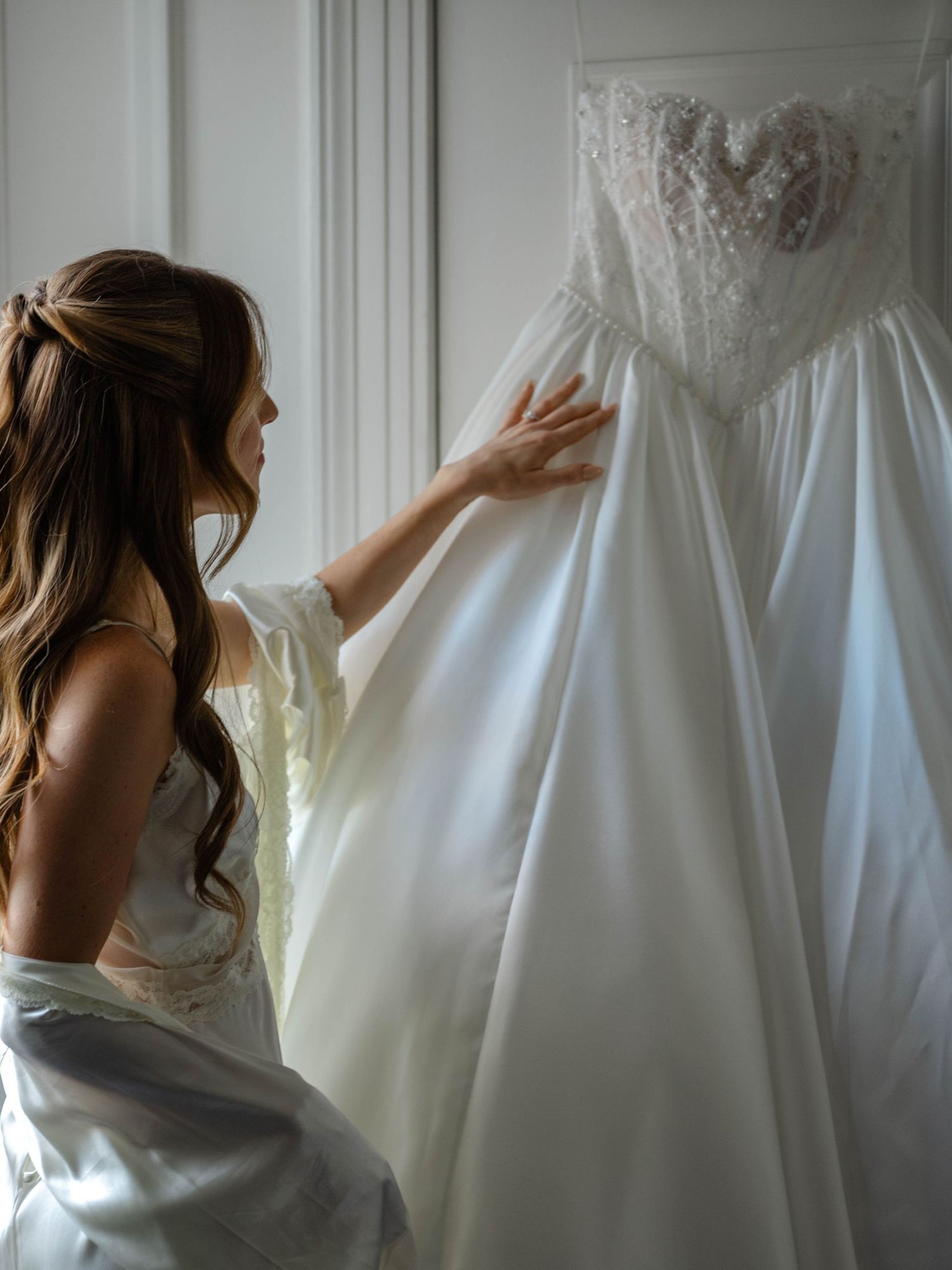 Bride with long wavy hair gently touches lace bodice of her strapless white ballgown wedding dress.