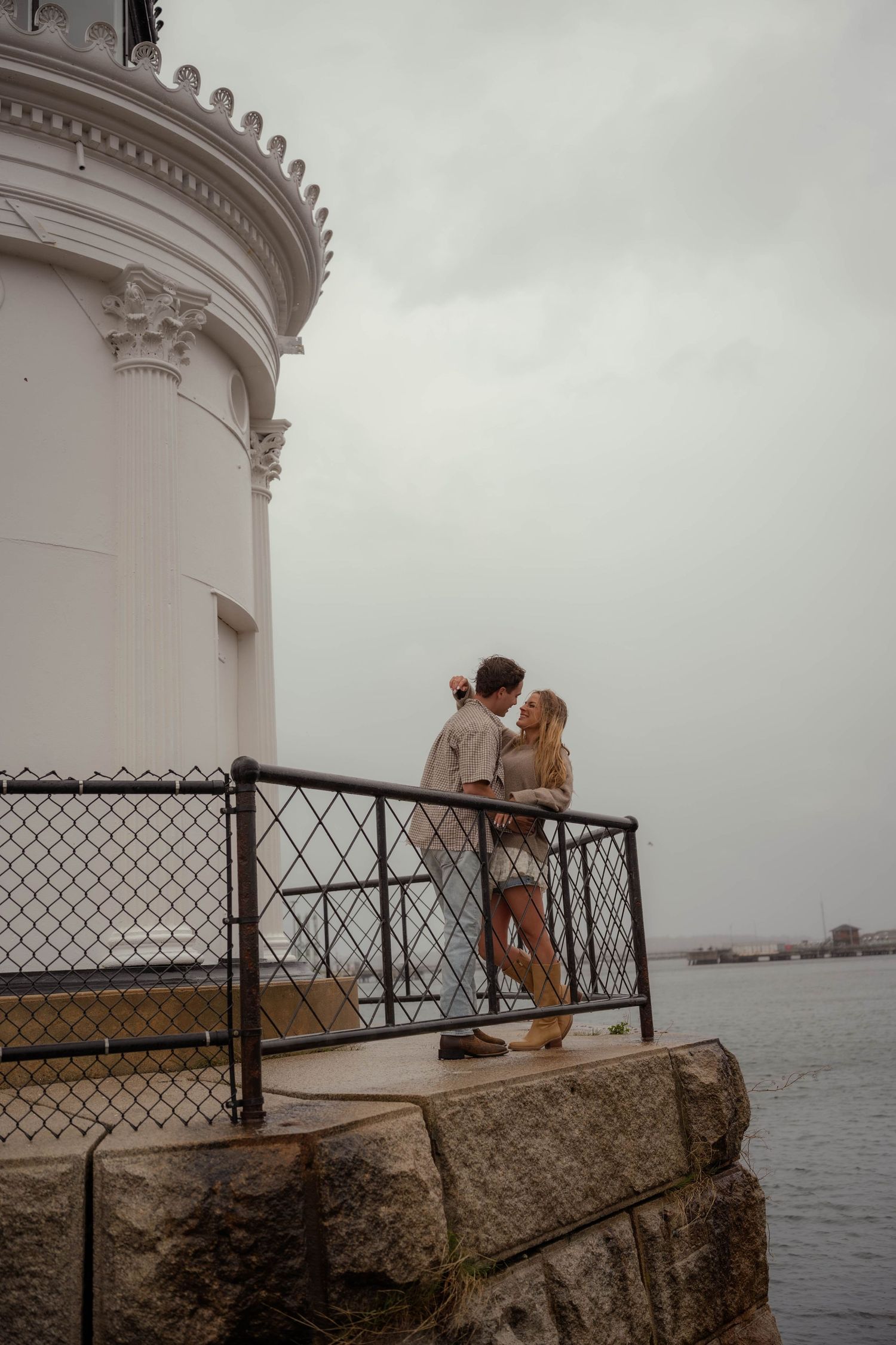 Couple sharing tender moment on rocky ledge beside white lighthouse by the sea.