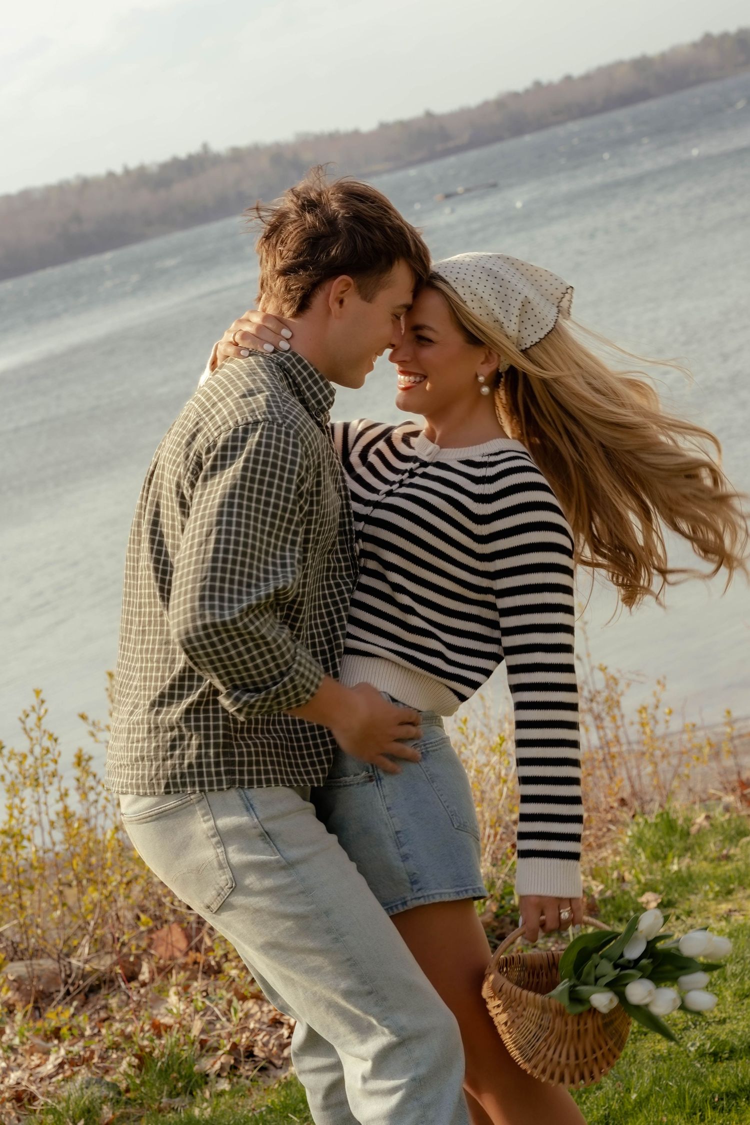 Happy couple nearly kissing outdoors by a lake, woman holding a basket of white tulips.